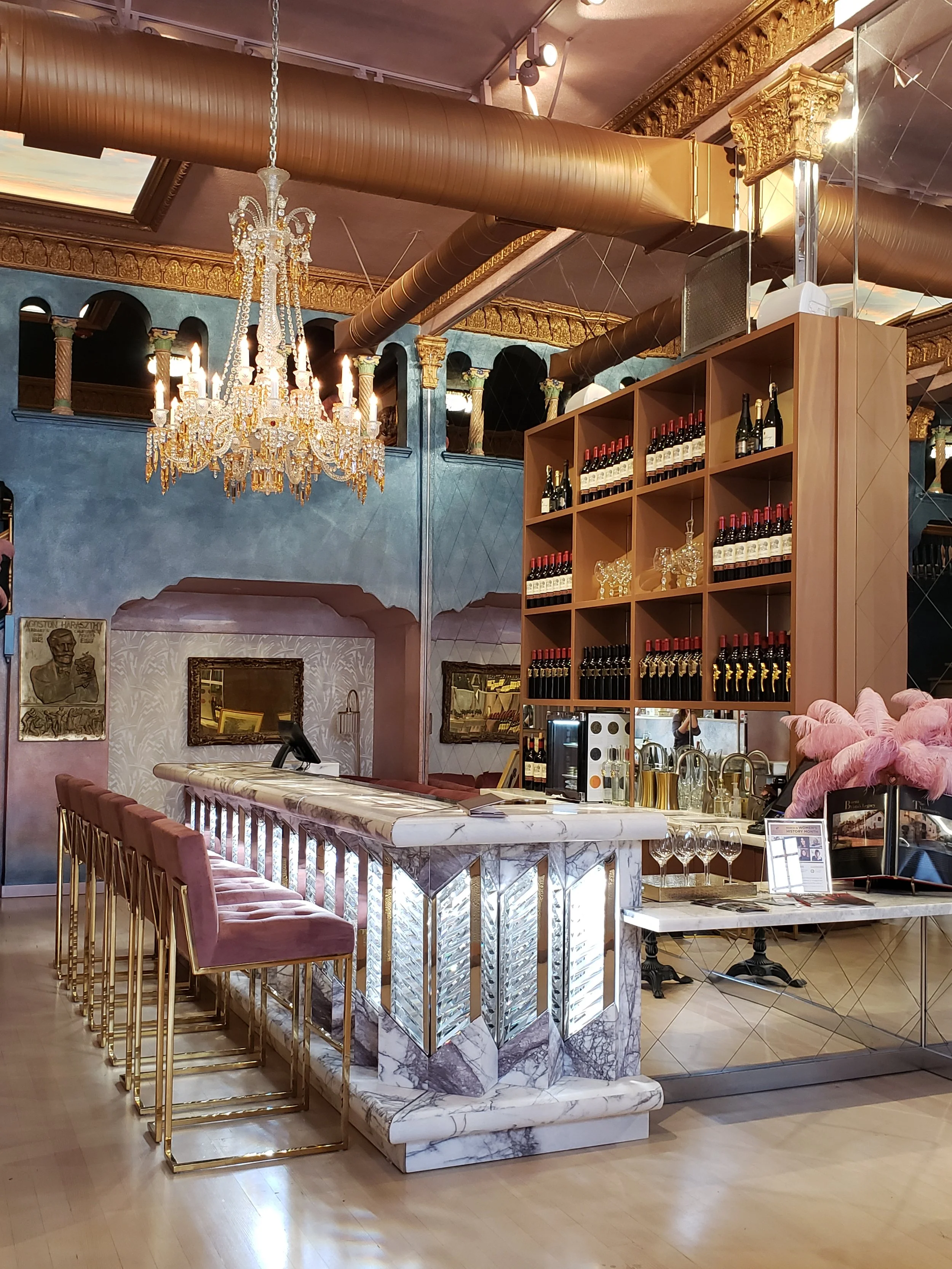 An elegant bar with a marble counter, pink velvet chairs with gold legs, a chandelier, and a wooden shelf filled with wine bottles.