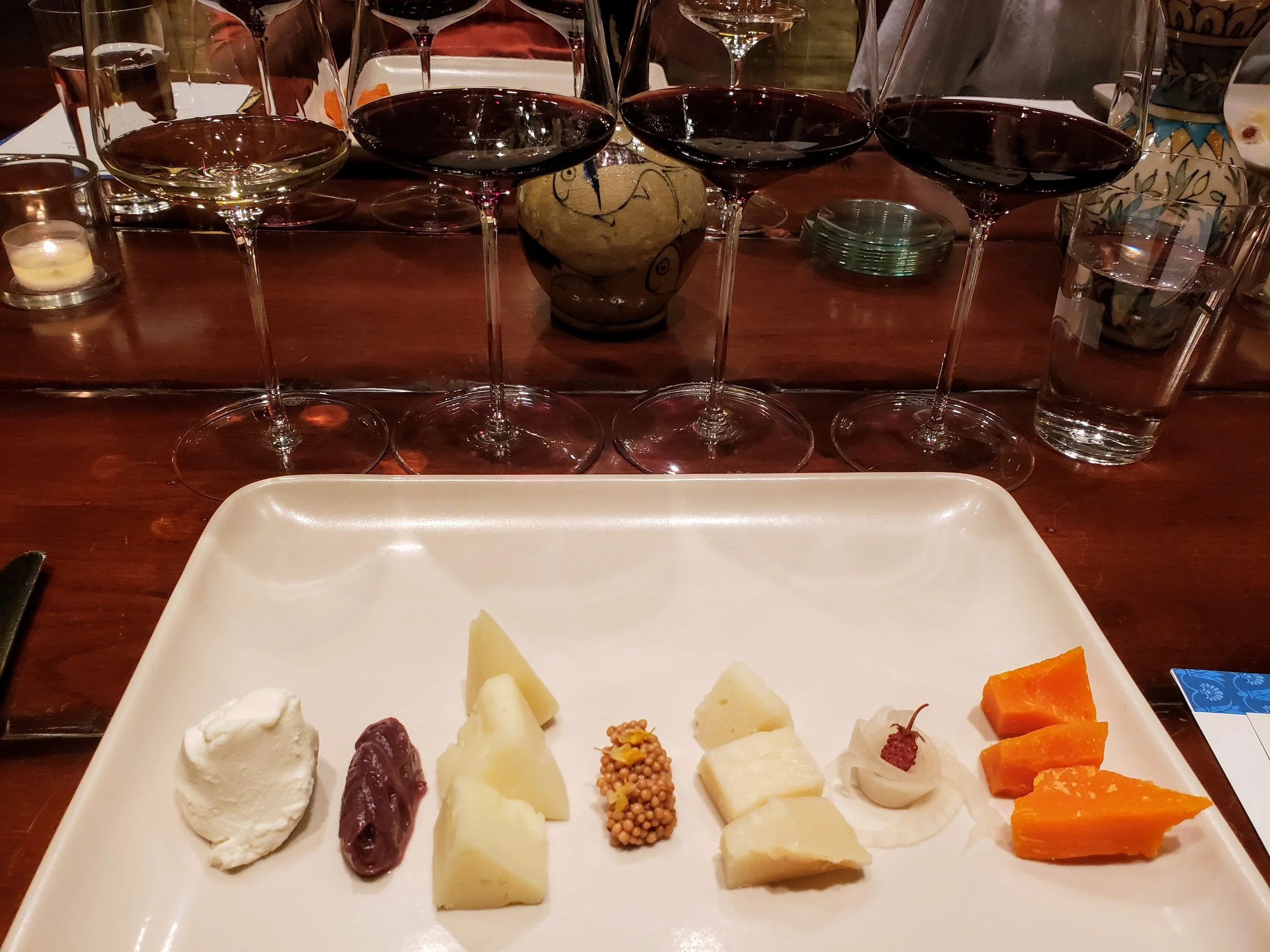 A cheese and dessert platter with various cheeses, a dollop of whipped cream with a cherry, slices of orange cheese, and blocks of hard cheese on a white plate. In the background, there are four glasses of red wine, two glasses of water, and candles 