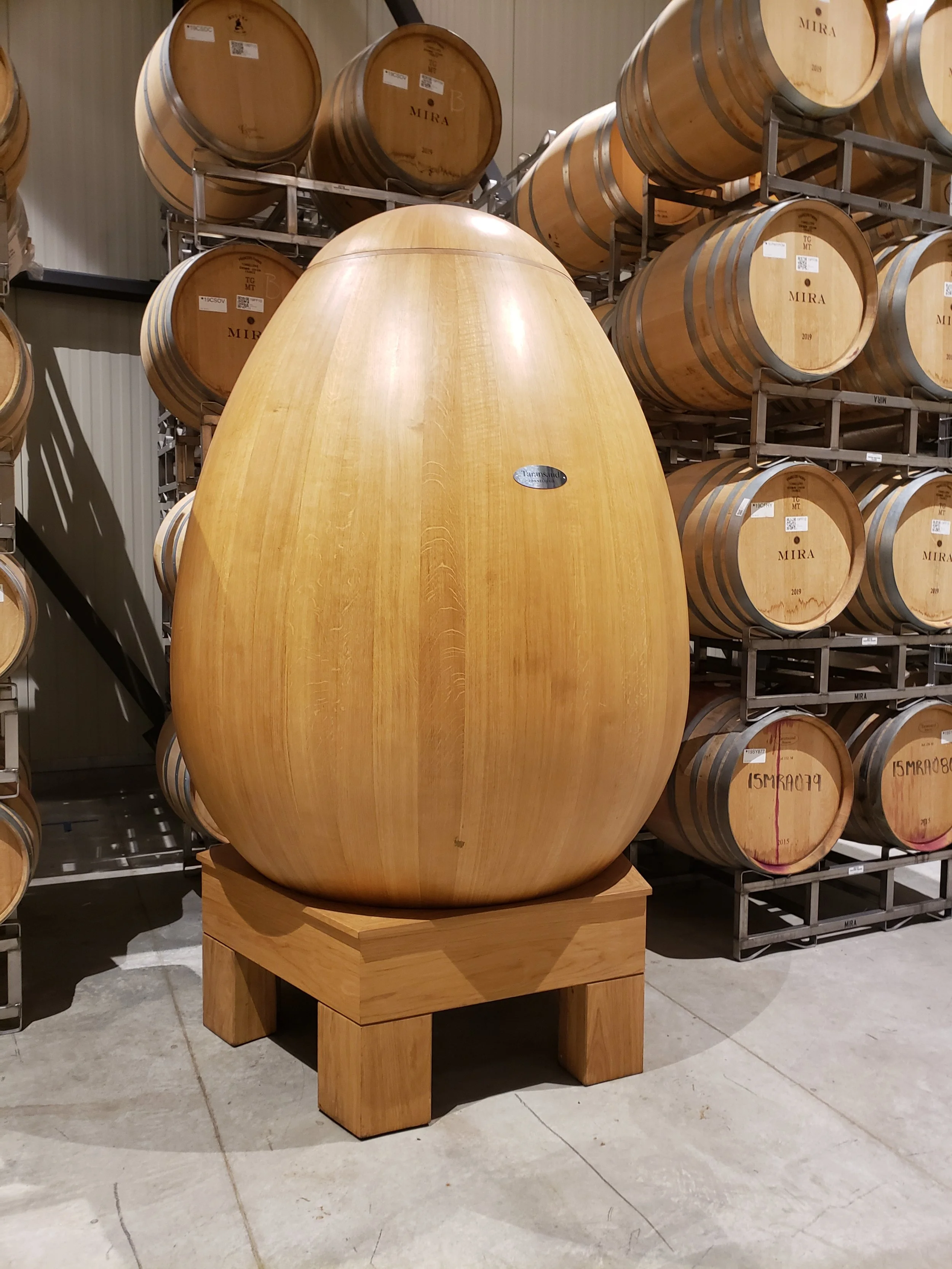 A large wooden egg for fermentation displayed on a wooden stand in a warehouse with wine barrels on shelves in the background.