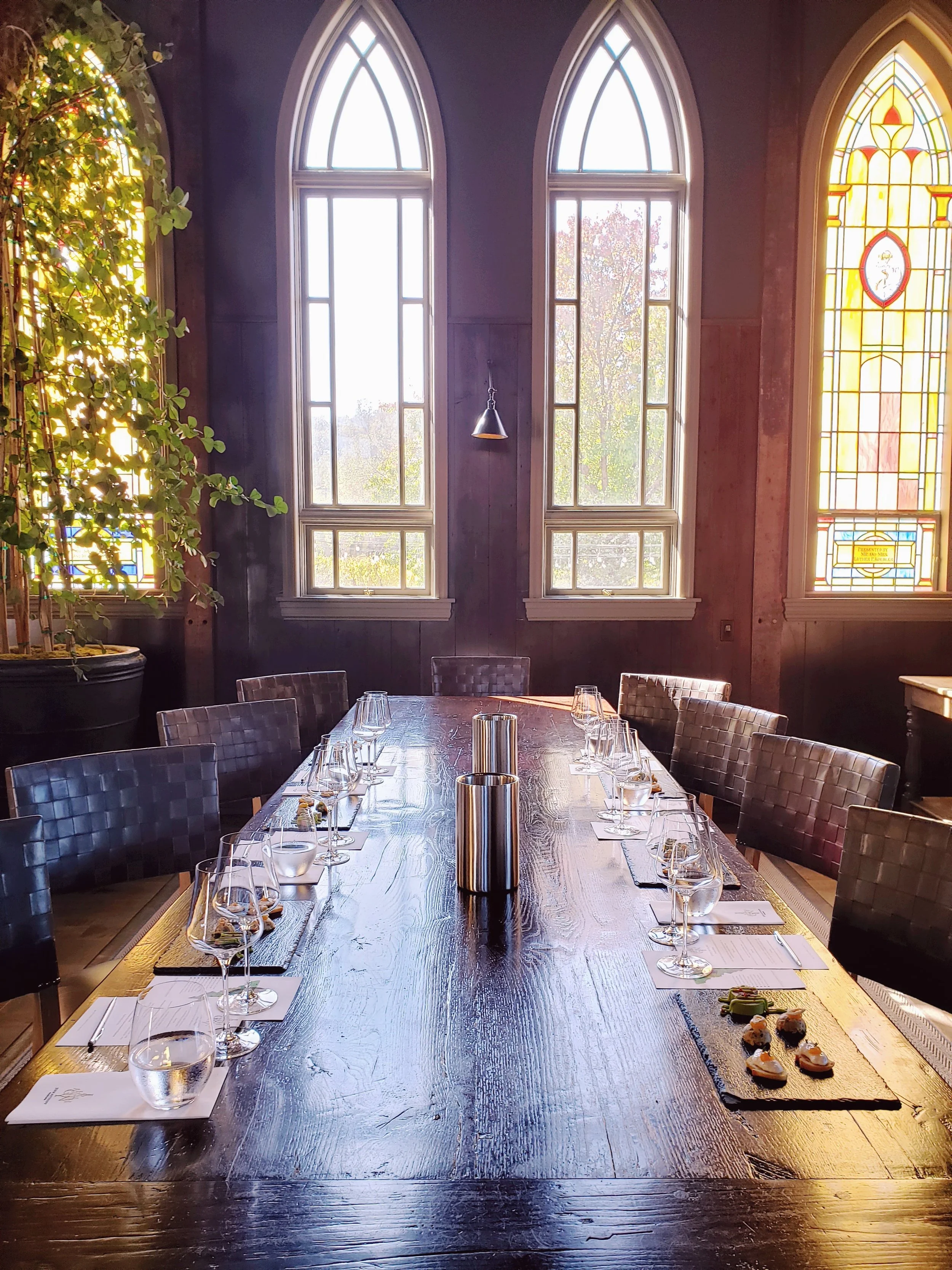 A dining room with a long wooden table set with water glasses, wine glasses, napkins, and small plates, in front of large arched windows with stained glass details. Sunlight streams through the windows, illuminating the table and dark wood-paneled wa