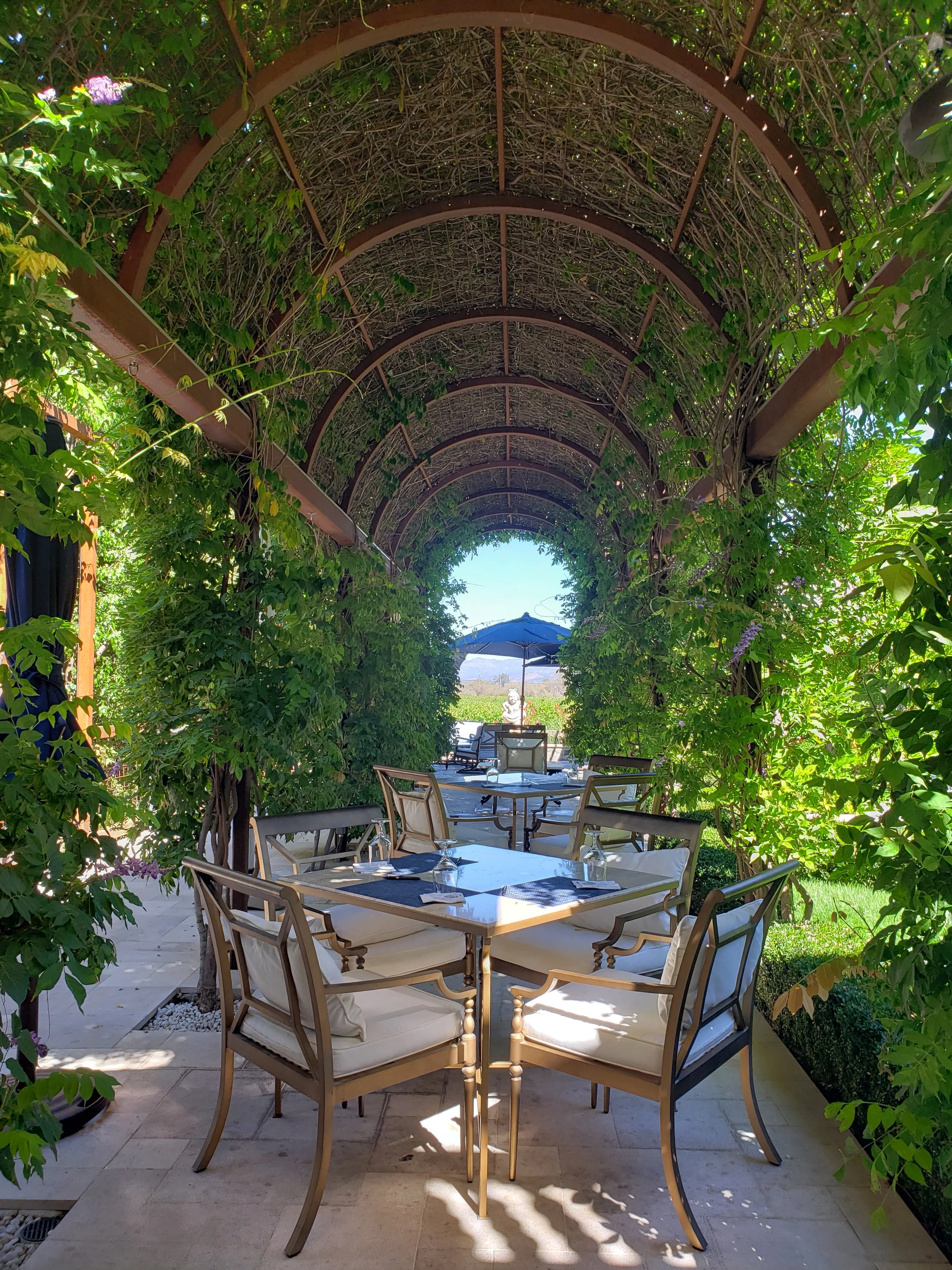 Outdoor dining area under a trellis with greenery and purple flowers, set with tables and chairs, shaded by a large umbrella in the background.