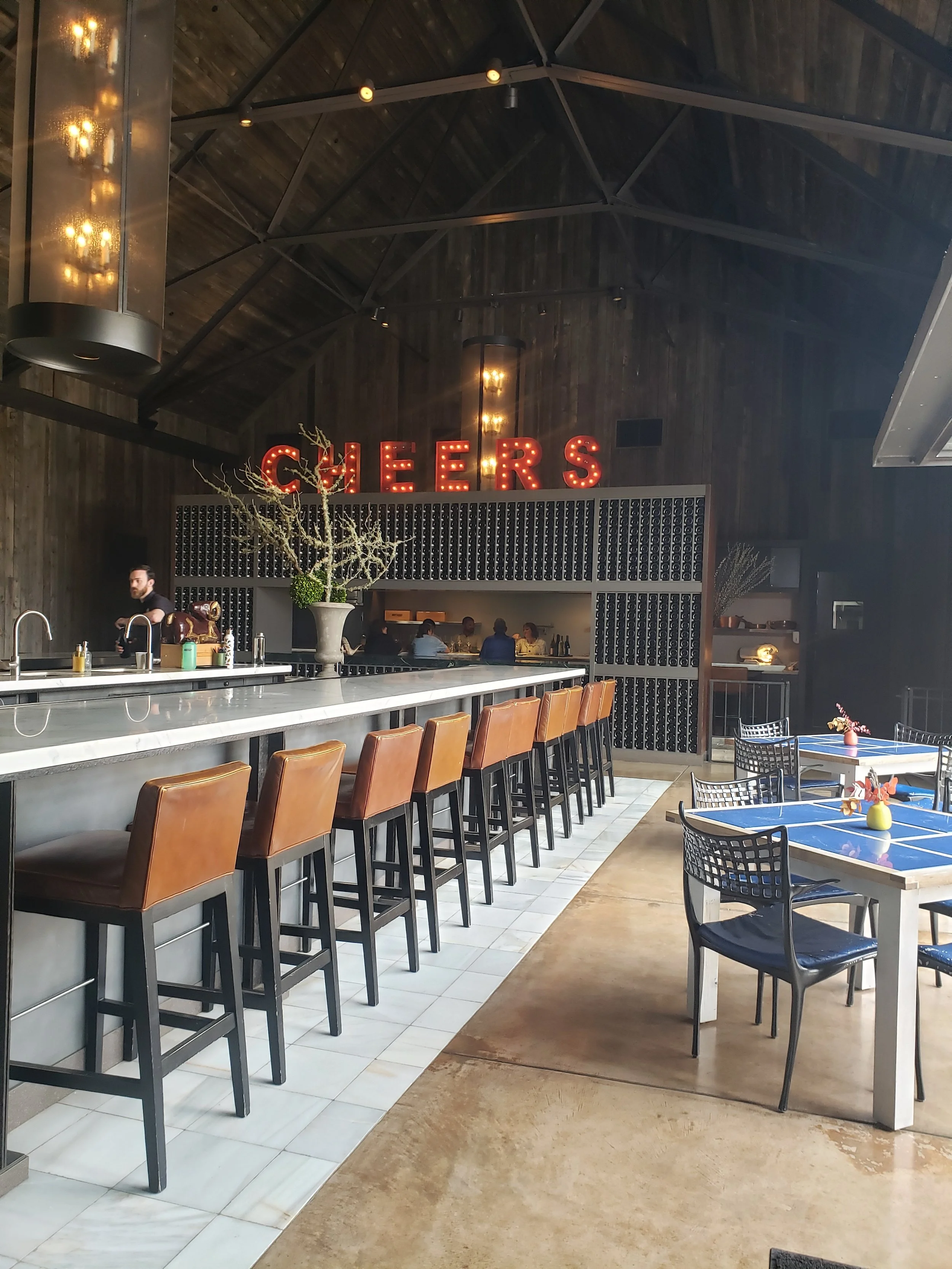 Interior of a modern restaurant bar with a row of high leather bar stools, a counter, and tables with chairs. Neon sign spelling 'CHEERS' hangs on the wooden wall.