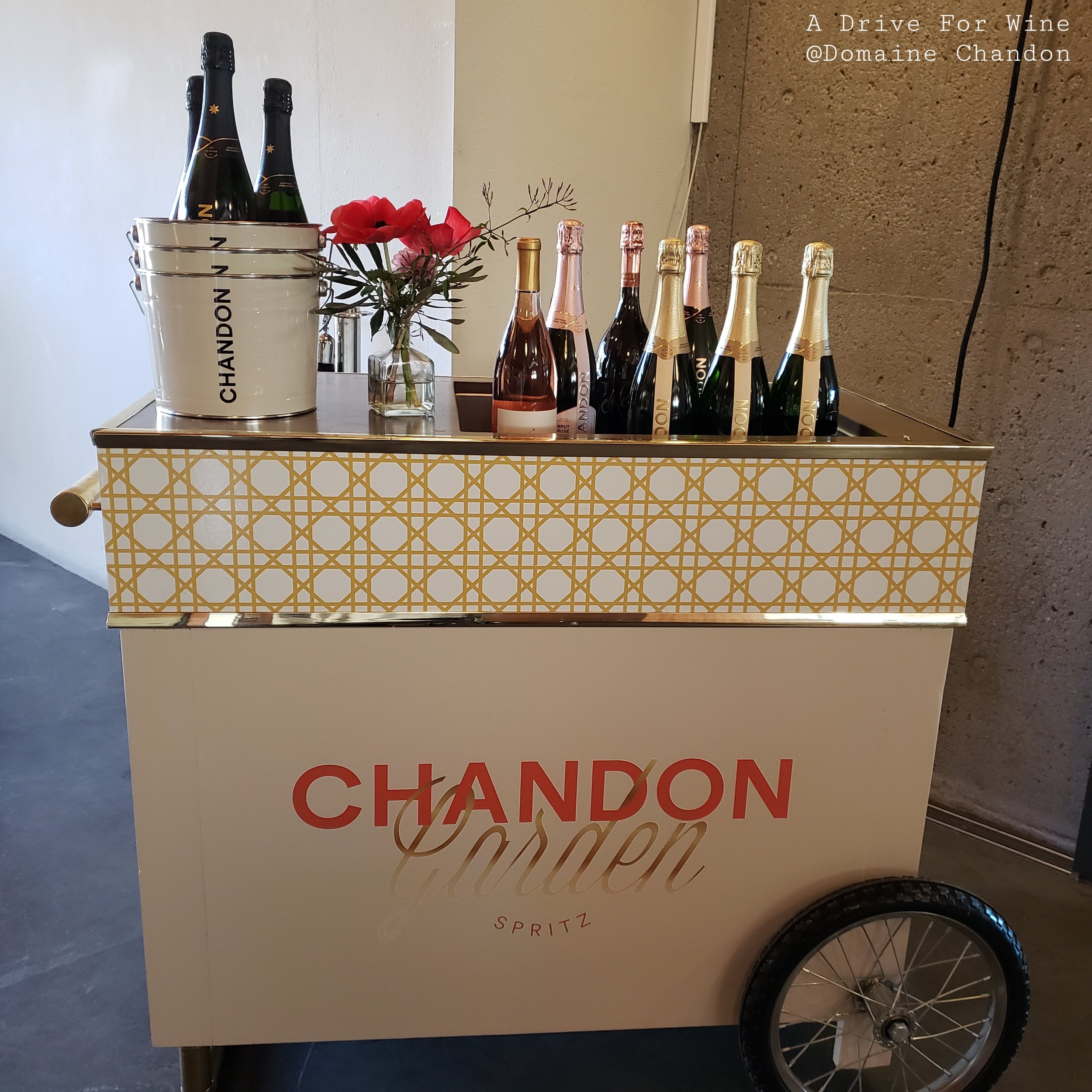 A mobile cart with a Chandon logo, containing bottles of sparkling wine, a small bouquet of flowers, and a large white ice bucket with Chandon branding.