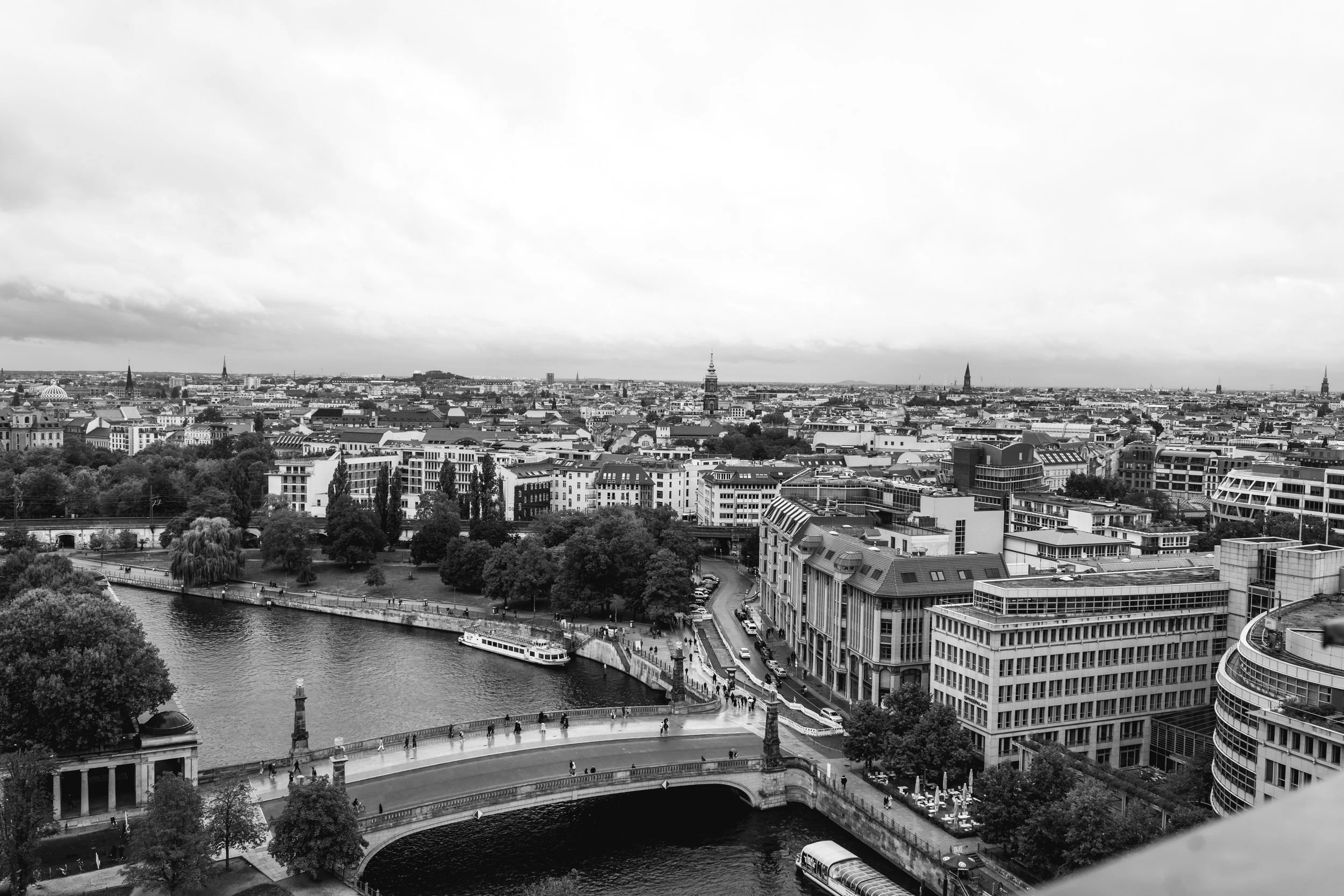 Black and white aerial view of a cityscape with a river, bridge, modern buildings, trees, and several church spires and towers in the background.