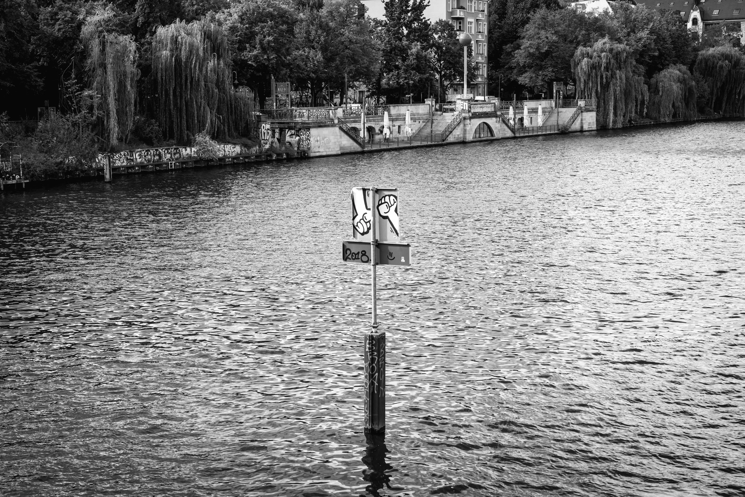 A pole in the water with graffiti art on a sign at a riverside park, trees, buildings, and a walkway in the background, all in black and white.