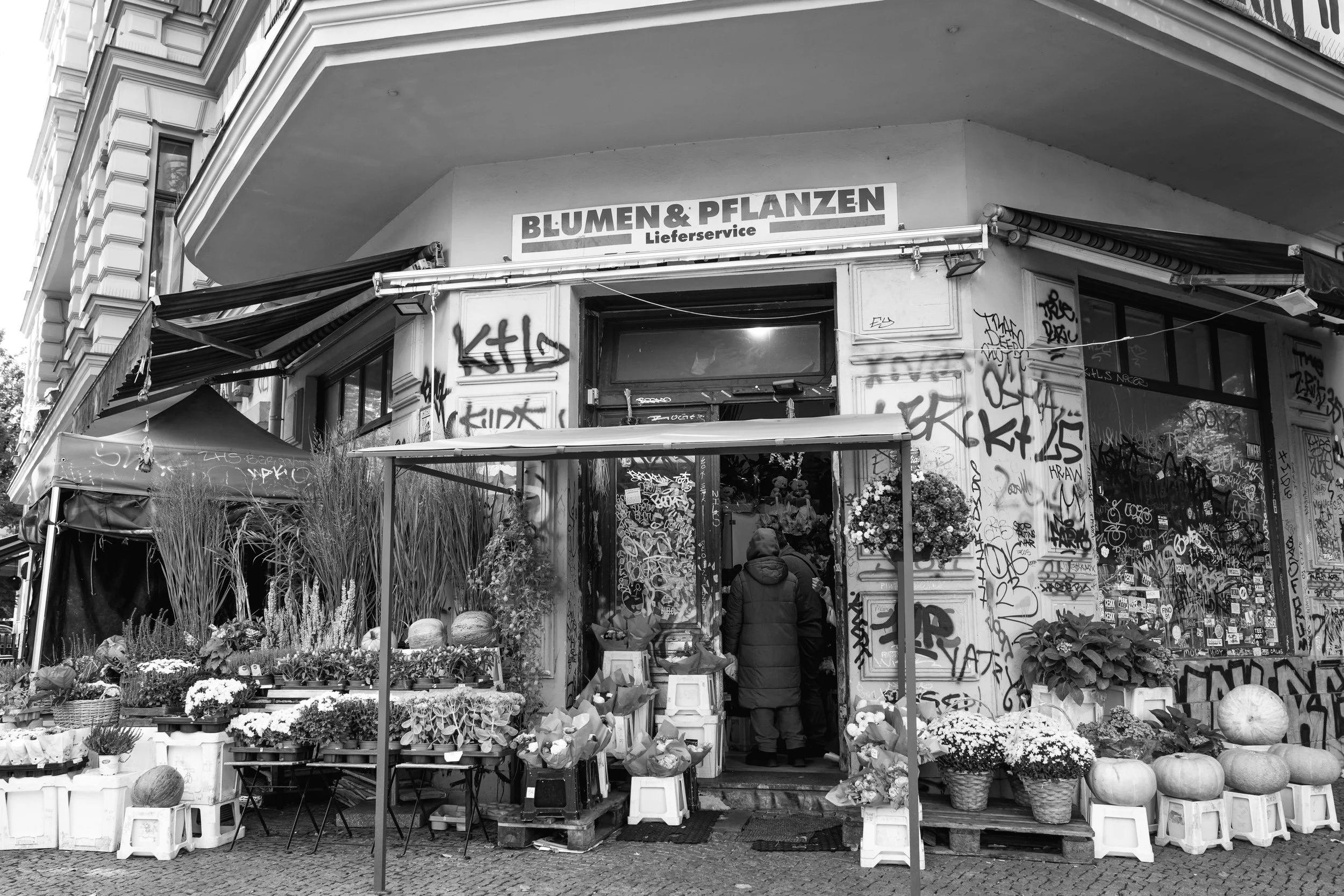 A flower shop with colorful plants and flowers outside, graffiti on the walls, and a sign that reads 'Blumen & Pflanzen Lieferservice'.