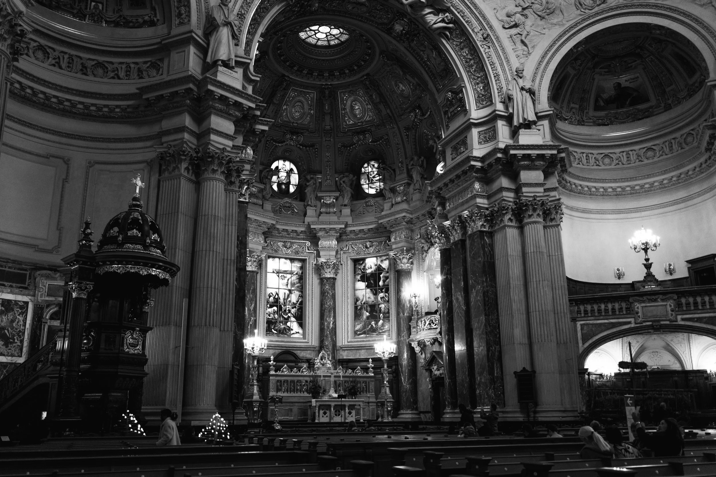 Interior of a grand church with ornate architecture, statues, stained glass windows, and a ceiling with detailed artwork, with people seated in pews.