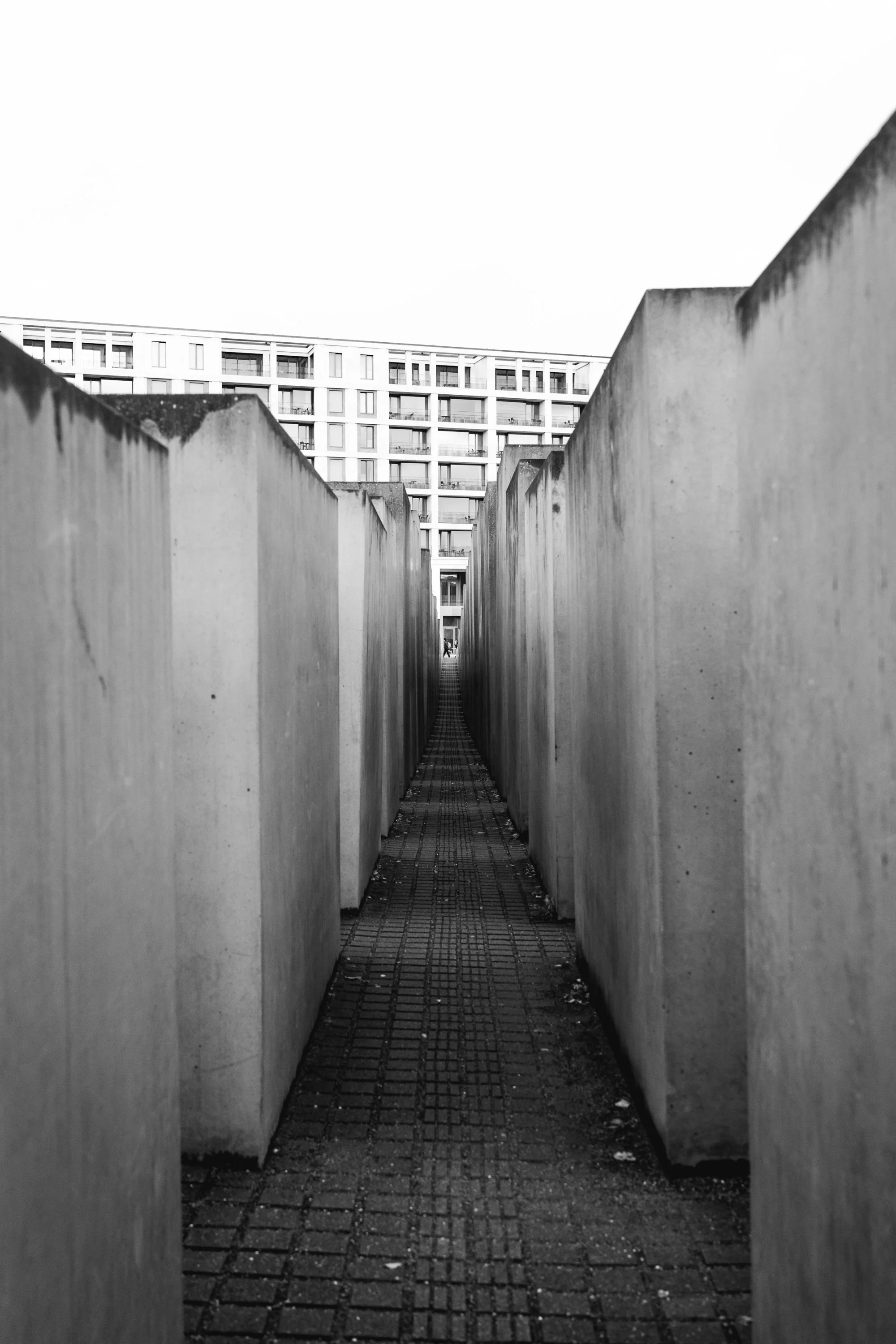 Black and white photo of the memorial to the victims of the Holocaust, consisting of concrete blocks arranged in a grid with a narrow pathway through the center, leading towards a building in the background.