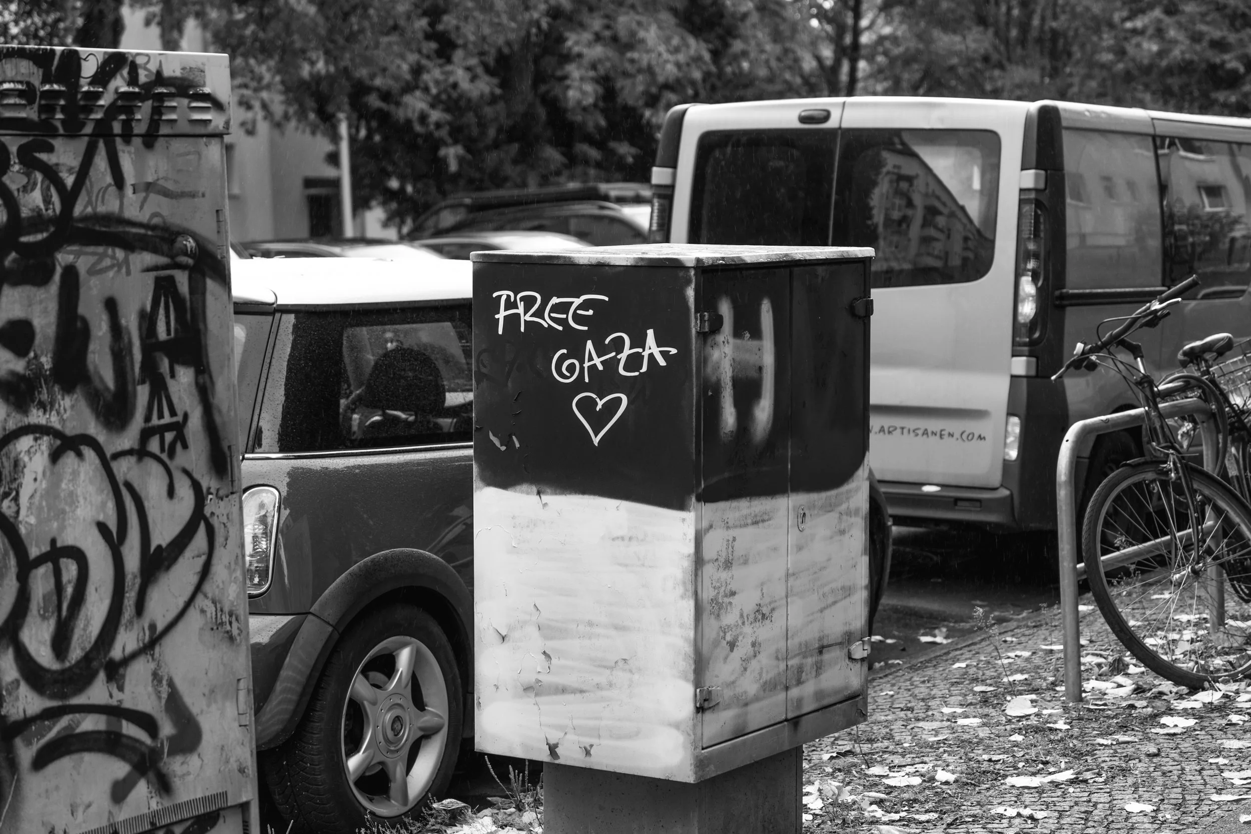 A street scene with graffiti-covered electrical box, a car, bicycles locked to a bike rack, and a van in the background. The electrical box has graffiti and the words 'Free Gaza' with a heart symbol written on it.