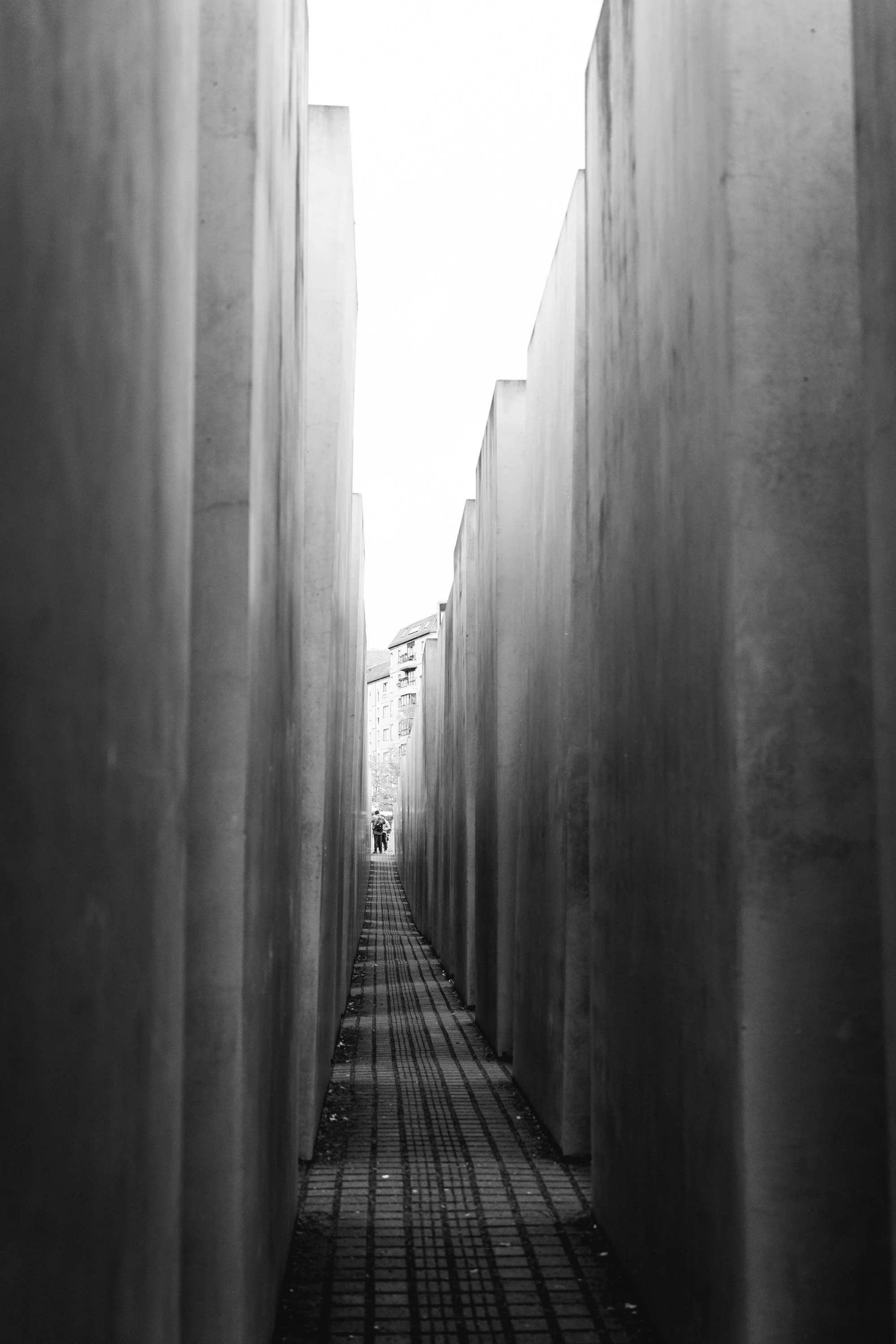 Black and white photo of narrow walkway between tall concrete slabs, with two people and buildings visible in the distance.