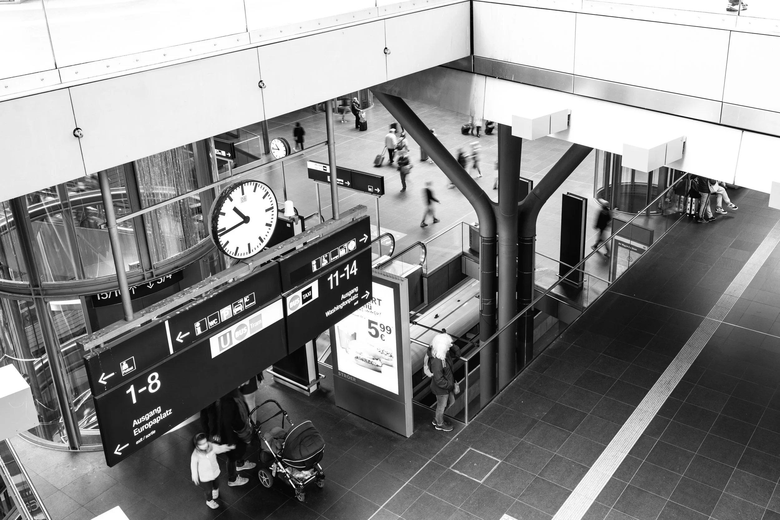 Interior view of a busy train station with signs, clocks, escalator, and people walking with luggage.