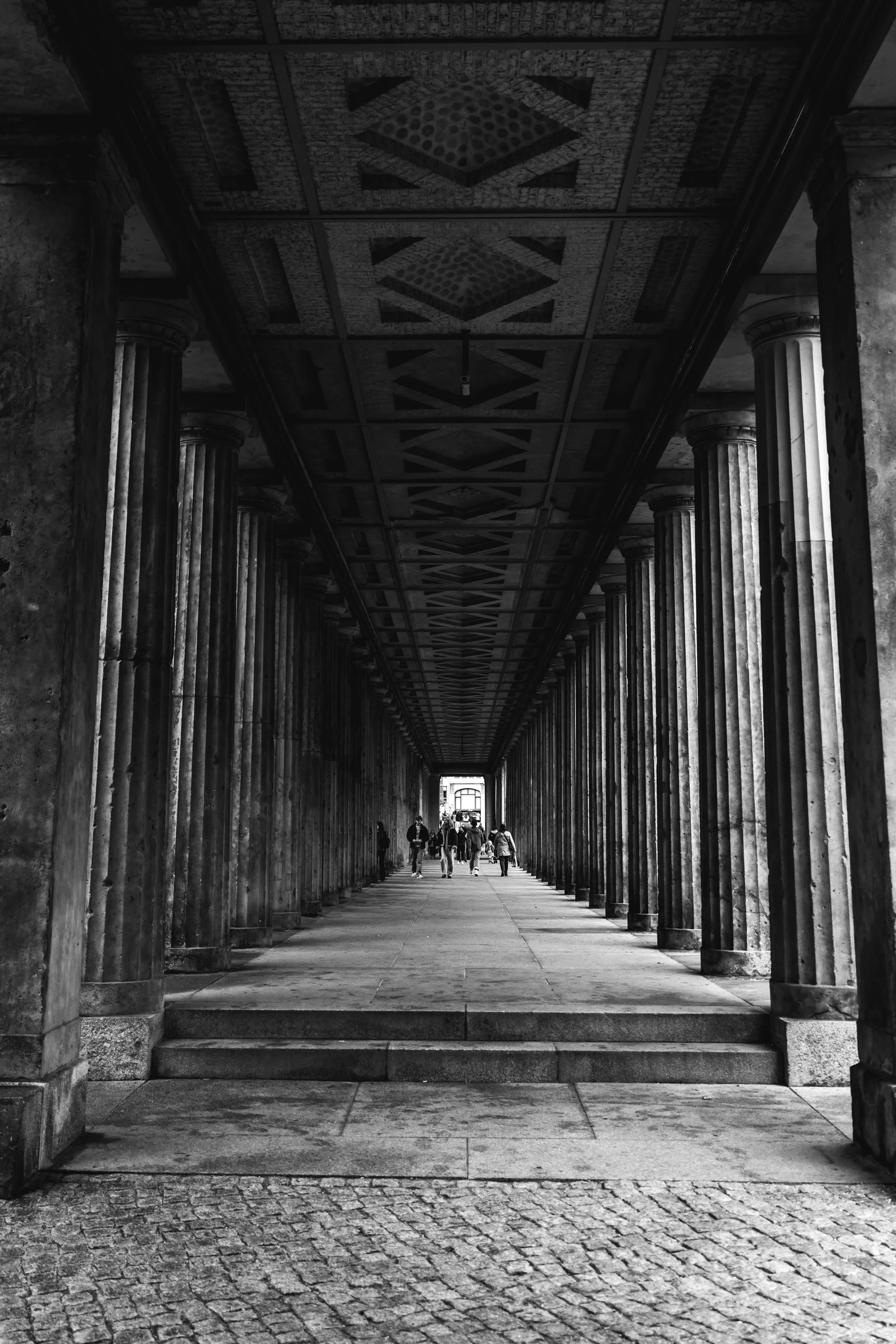 Black and white photo of a colonnade with tall columns and a patterned ceiling, with people walking towards an illuminated opening at the end.