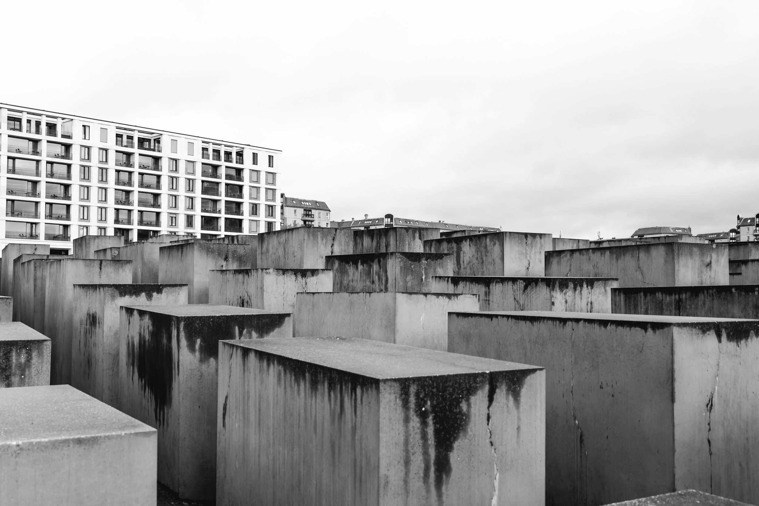 Black and white photo of concrete blocks in the Holocaust Memorial in Berlin with a modern apartment building in the background under cloudy sky.
