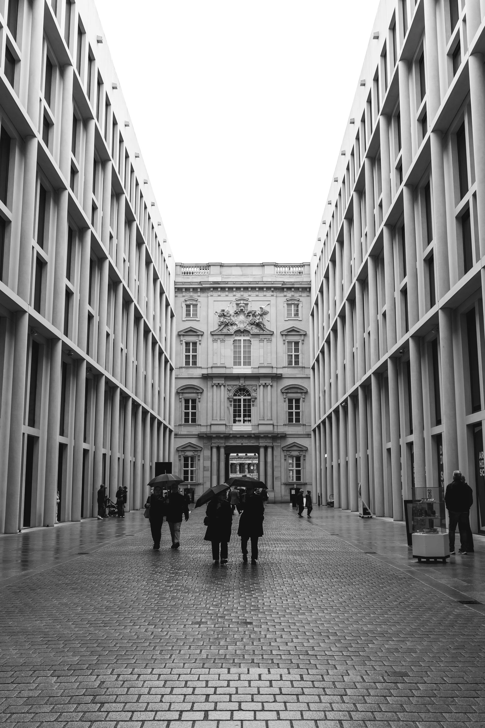 People walking with umbrellas in a modern urban plaza flanked by tall, symmetrical buildings, leading to an ornate, historic building in the background in black and white.
