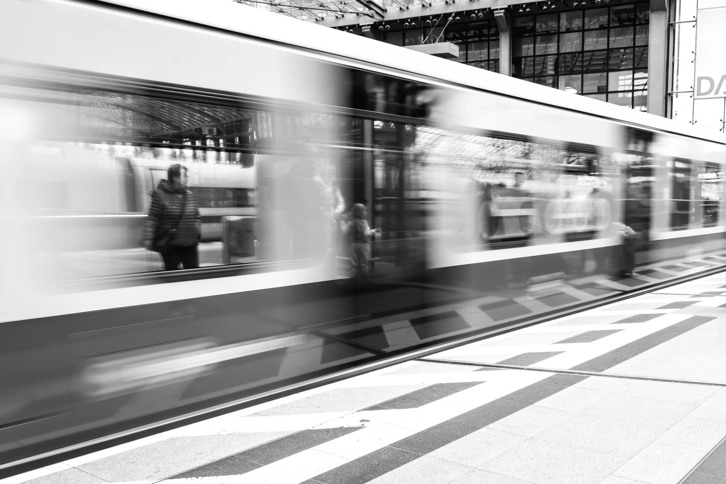 Blurred image of a train at a station with a person waiting on the platform, reflected in the train windows, in black and white.