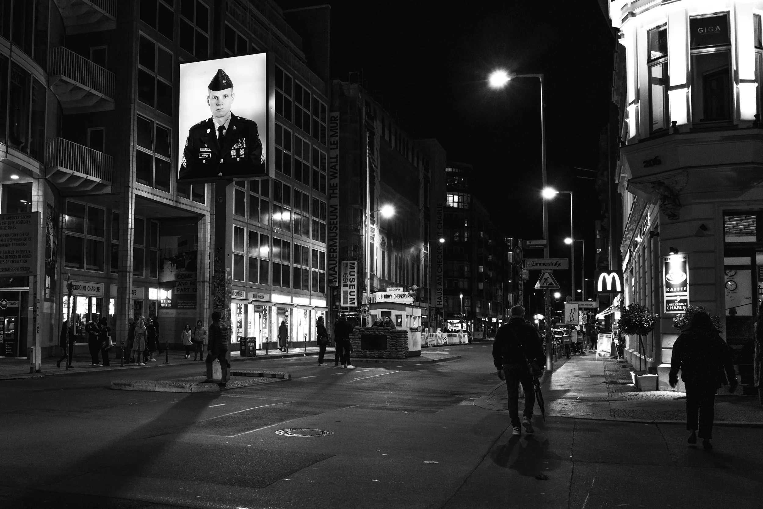 A black and white photo of Checkpoint Charlie at night, with pedestrians walking and various illuminated signs, including a McDonald's logo and a large billboard featuring a person in military uniform.