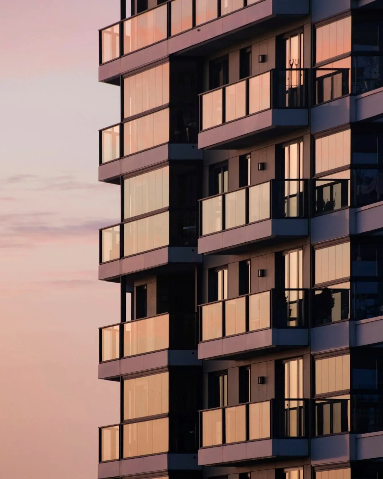 A modern multi-story building with glass balconies reflects the pinks and oranges of a sunset sky.