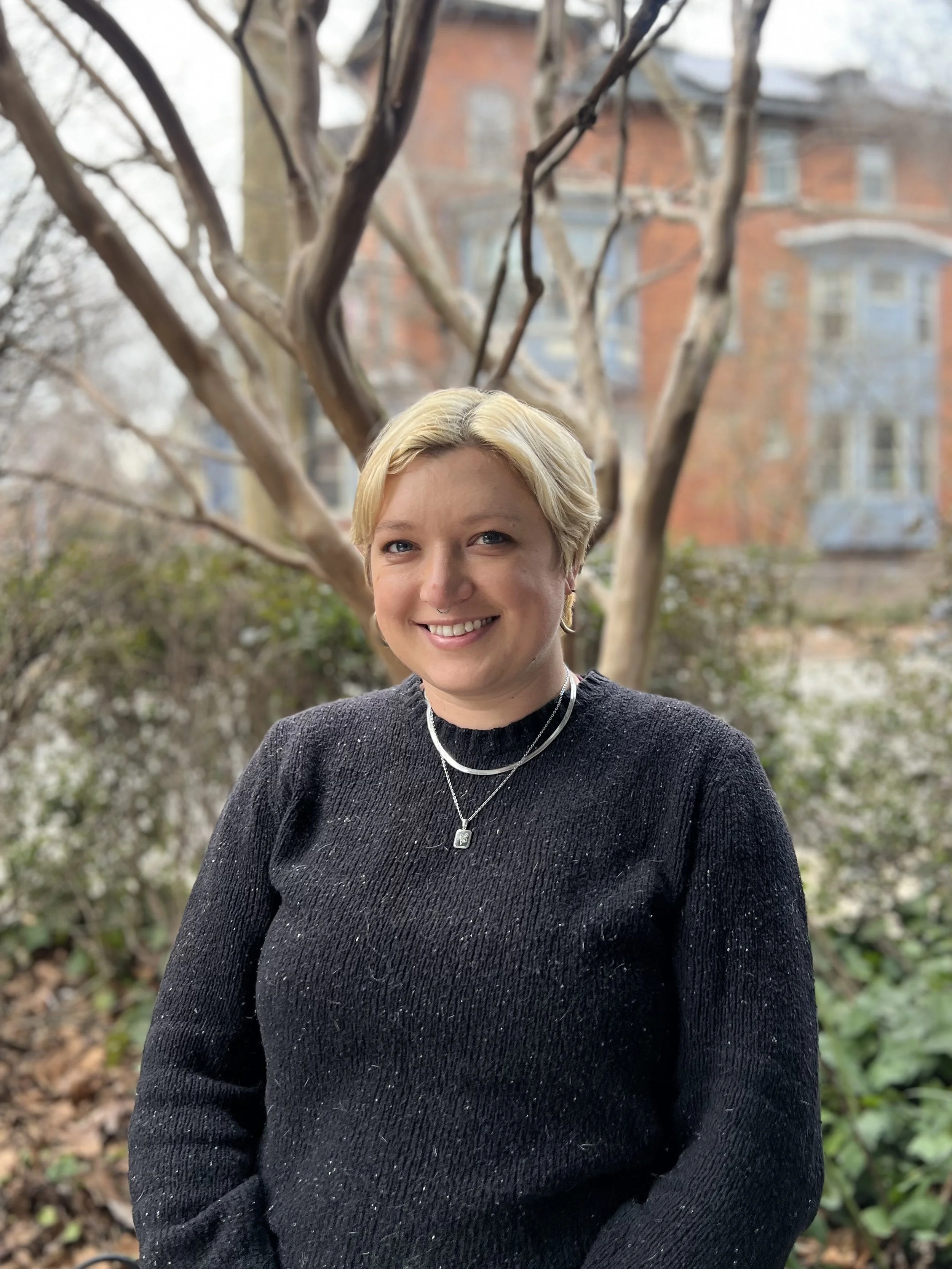 Person smiling outdoors in front of a brick building and trees, wearing a black sweater and necklace.