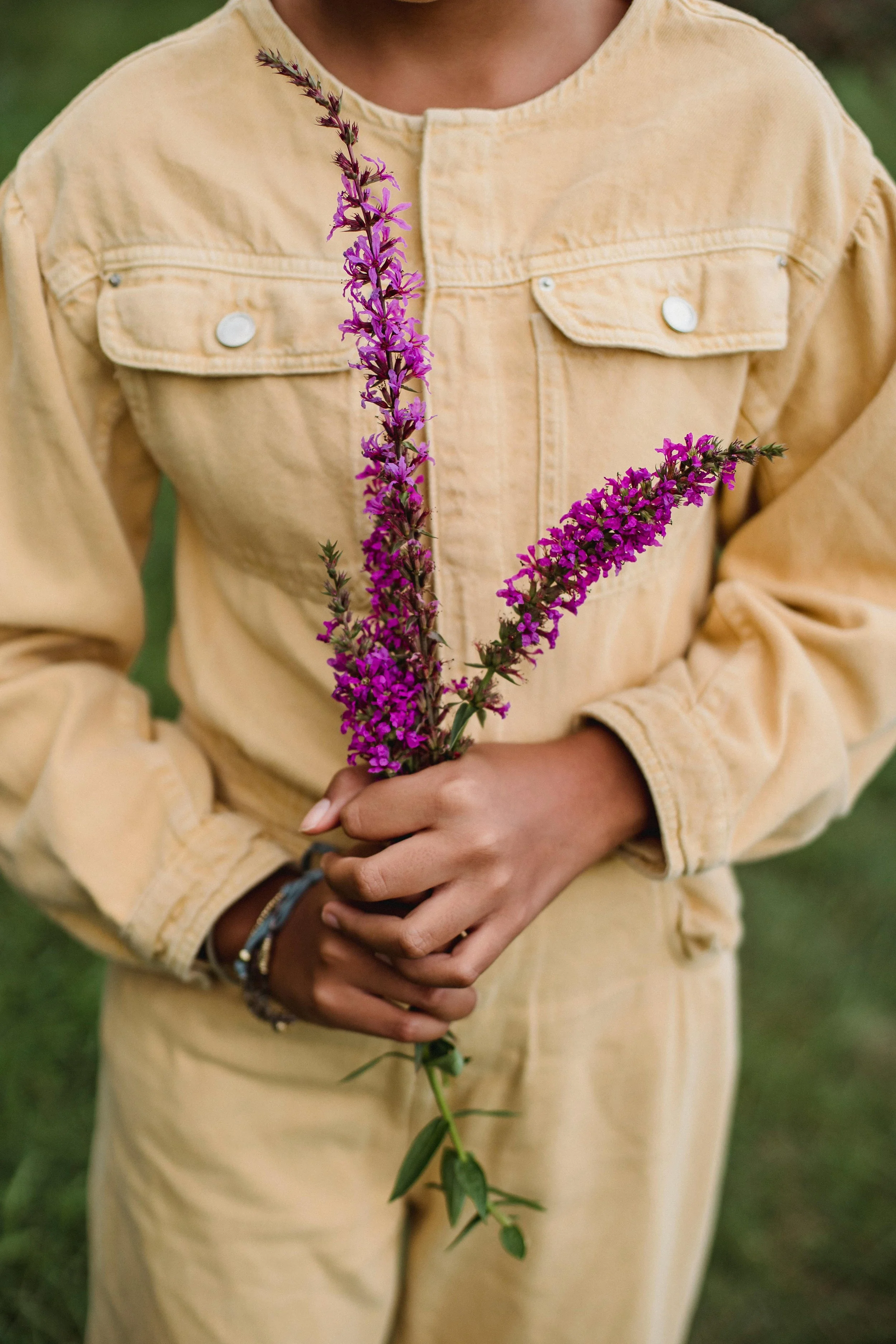 Person in a beige jumpsuit holding purple flowers outdoors.