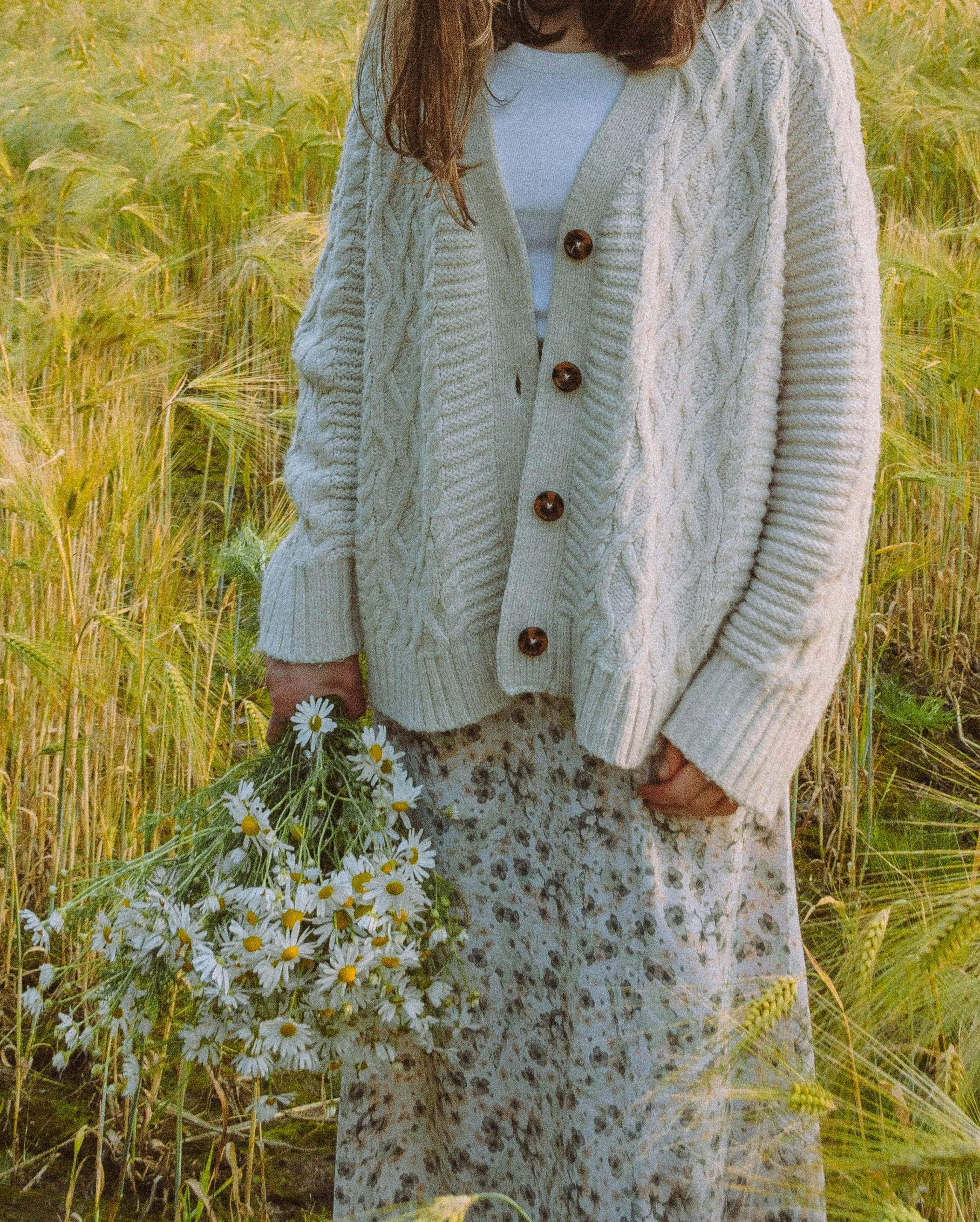 Woman in a beige cardigan and floral skirt holding a bouquet of daisies, standing in a field of tall grass.
