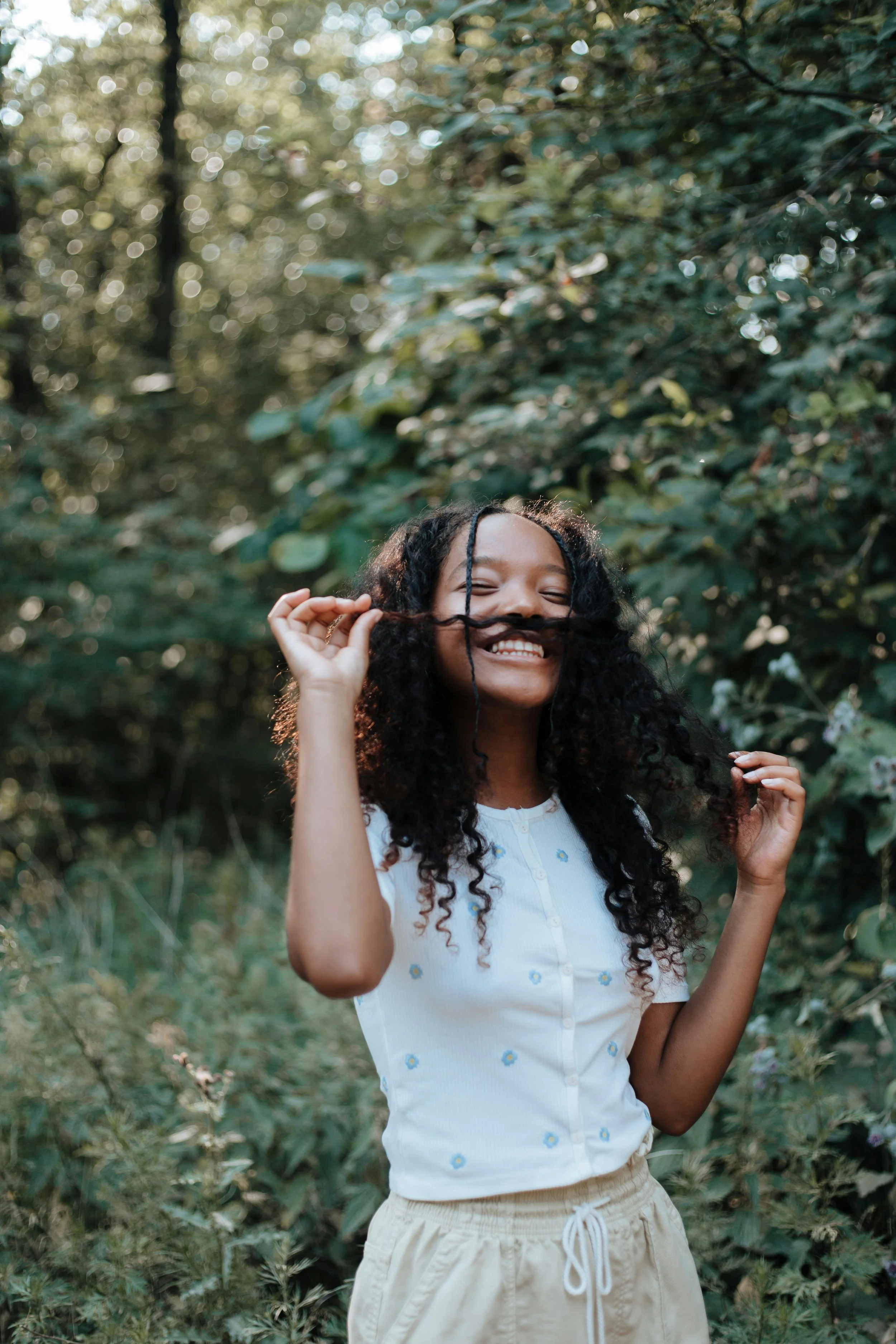 Person with curly hair holding a strand as a pretend mustache, smiling in a forest setting.