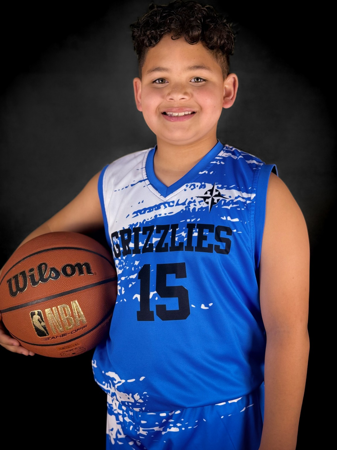 A young boy in a blue Grizzlies basketball uniform holding a basketball, smiling at the camera on a black background.