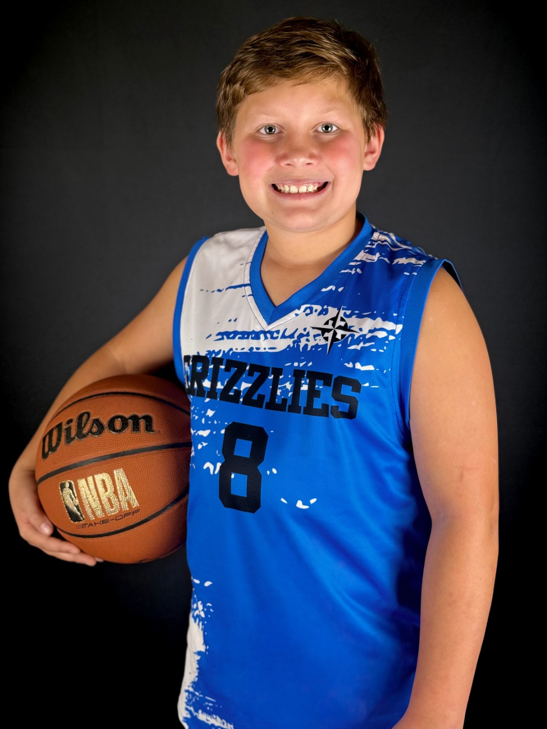 Young boy wearing a blue Memphis Grizzlies basketball jersey with the number 8, holding a Wilson NBA basketball, smiling, against a black background.