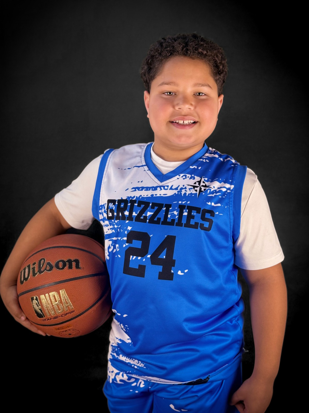 A young boy in a blue basketball jersey with the number 24 and 'Grizzlies' on it, holding a basketball, standing against a black background, smiling.