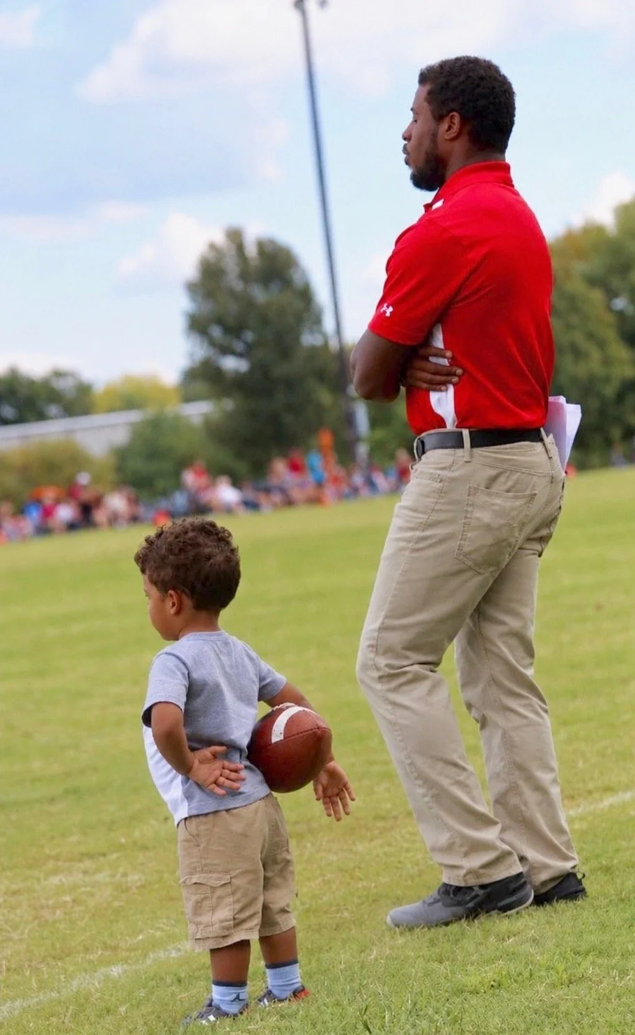 A young boy standing on a grassy field holding a football behind his back, while an adult man in a red polo shirt and beige pants stands nearby with his arms crossed, watching something off-frame. In the background, there are trees and a crowd of people sitting on the grass.