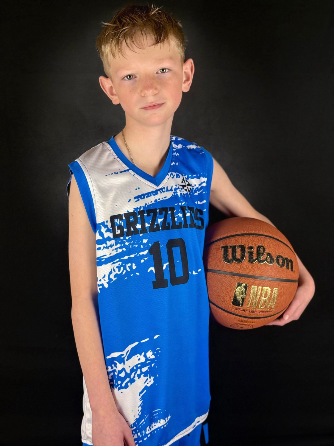 Young boy in a blue and white basketball uniform holding a Wilson NBA basketball, standing against a black background.