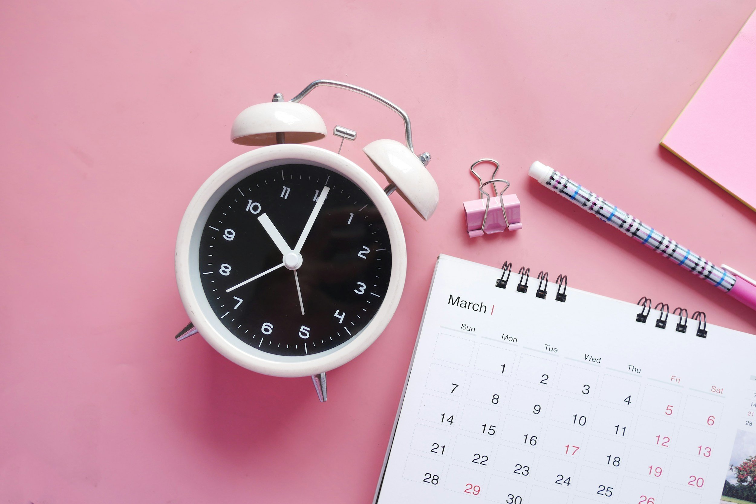 A white alarm clock showing 11:55, a pink binder clip, a white and pink patterned pen, and a March calendar on a pink background.
