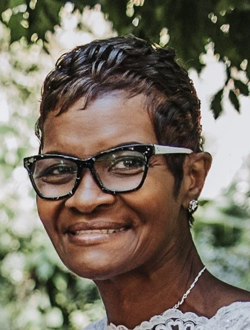 Smiling woman with short curly hair wearing black glasses, earrings, and a necklace outdoors with trees in the background.