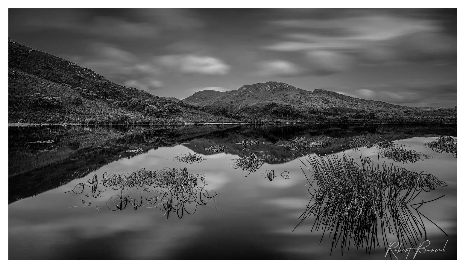 Black and white photograph of low hills and shoreline vegetation reflected in still water beneath a cloudy sky.
