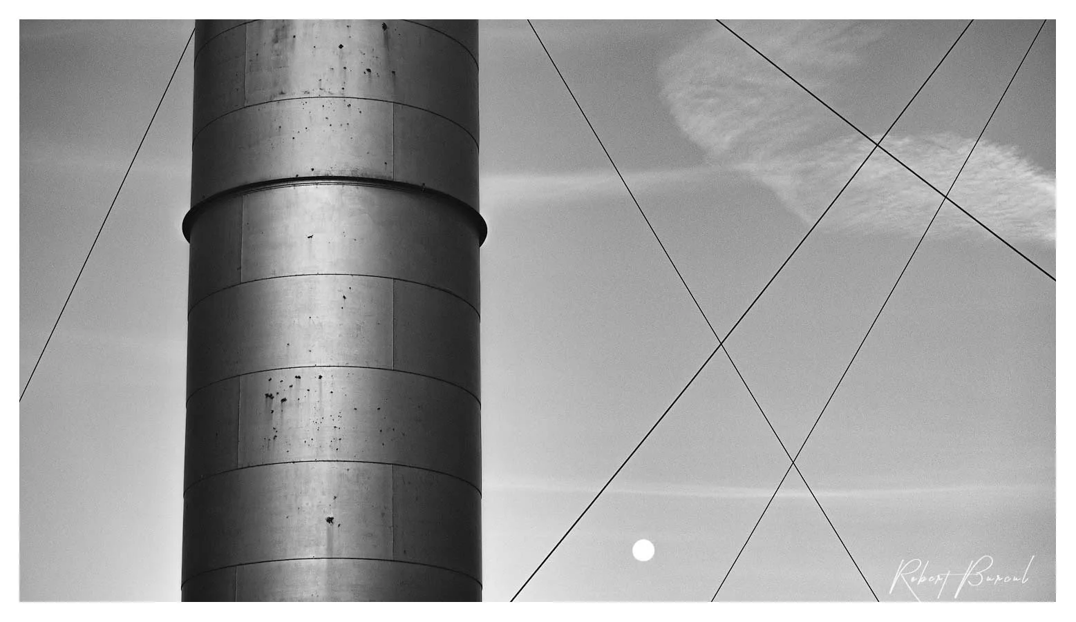 Black and white photograph of a cylindrical metal column structure with wires stretching as fine diagonal lines against the sky and faint moon shape.
