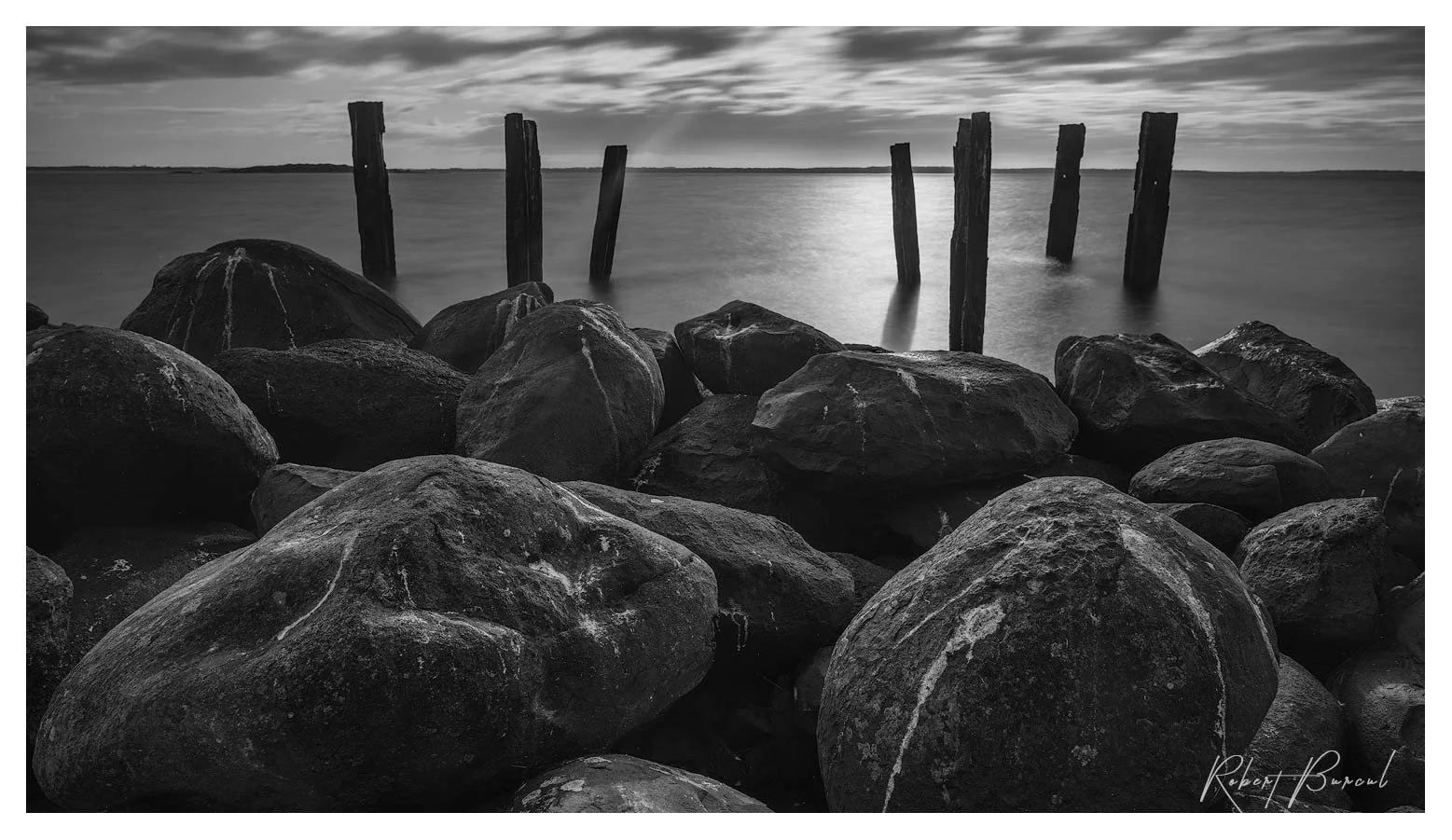 Black and white photograph of rounded shoreline rocks with weathered steel posts standing in the water beyond.