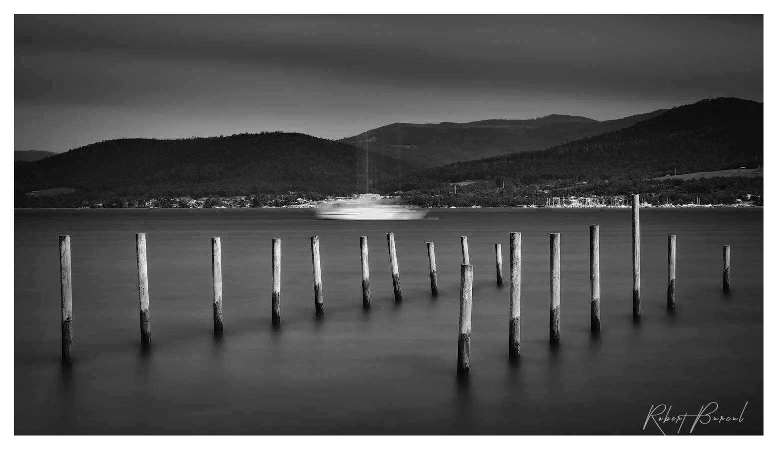 Black and white photograph of rows of timber posts rising from smooth water marking the boat ramp passage, with hills along the distant shore.