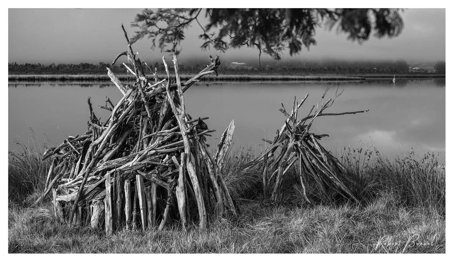 Black and white photograph of branches gathered into a rough pyramid structure on a grassy bank beside the water.