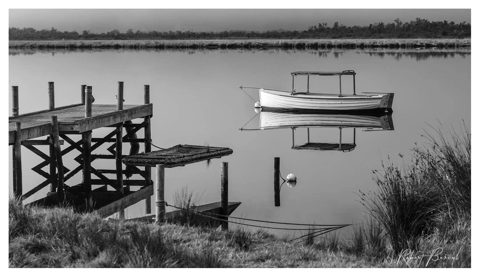 Black and white photograph of a small boat floating beside a timber jetty , reflecting on calm river surface.