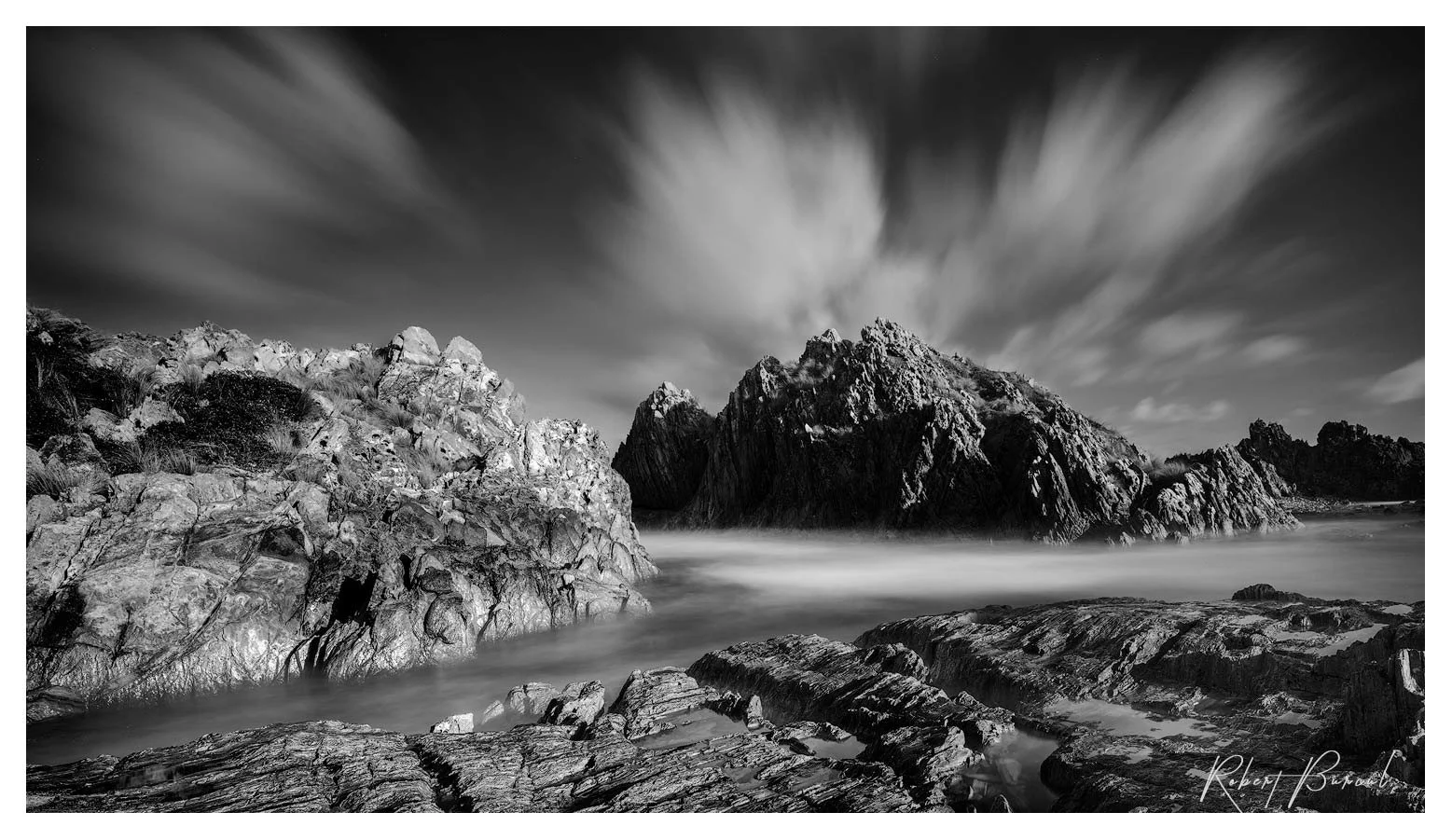 Black and white photograph of rugged rocks and rough water beneath streaked clouds.