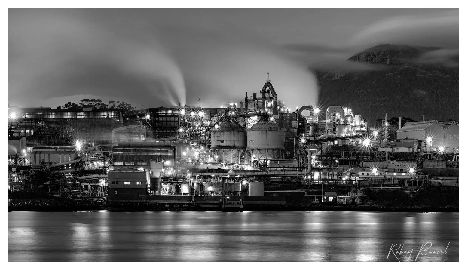 Black and white photograph of an illuminated industrial waterfront at night, with buildings and lights reflected in the water.