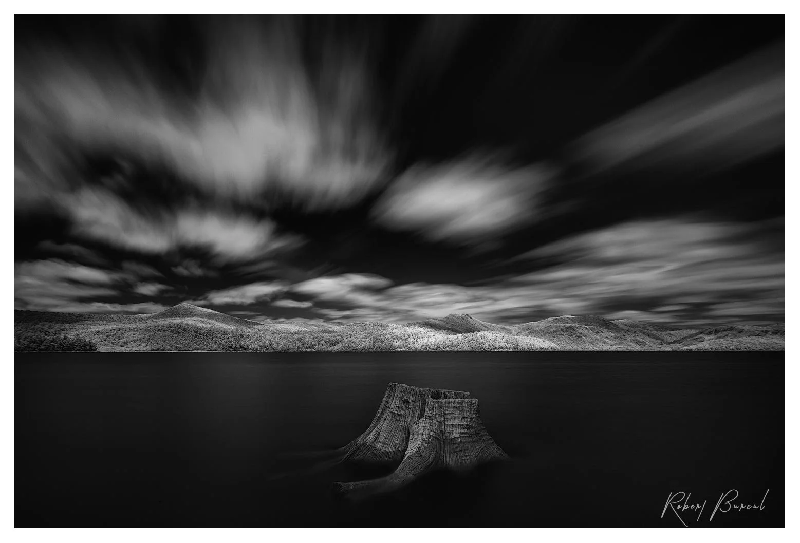 A long exposure infrared black and white image of lake with white streaking clouds and remains of a tree stump