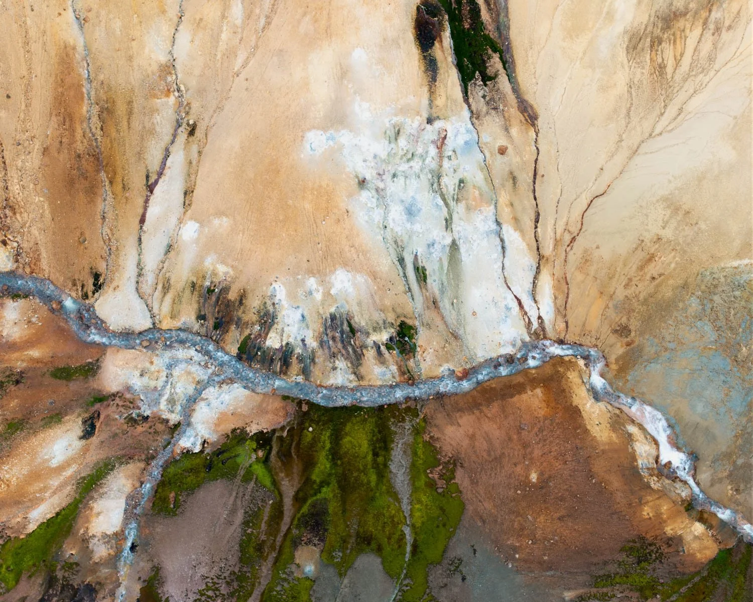 Geothermal Delta.  A geothermal spring cuts through mineral ground in Iceland — a sinuous blue-grey channel dividing warm sandy earth above from vivid emerald algae below, with extraordinary branching tributary forms. Dramatic, painterly large format