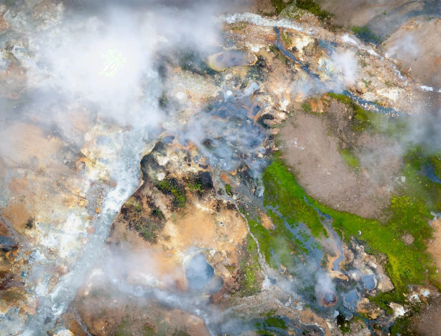Geothermal Steam.  A geothermal field in Iceland from above — springs billowing white steam revealing vivid green algae, amber mineral ground and blue-grey pools beneath. The earth, still becoming itself. Rare, atmospheric wall art for collectors and