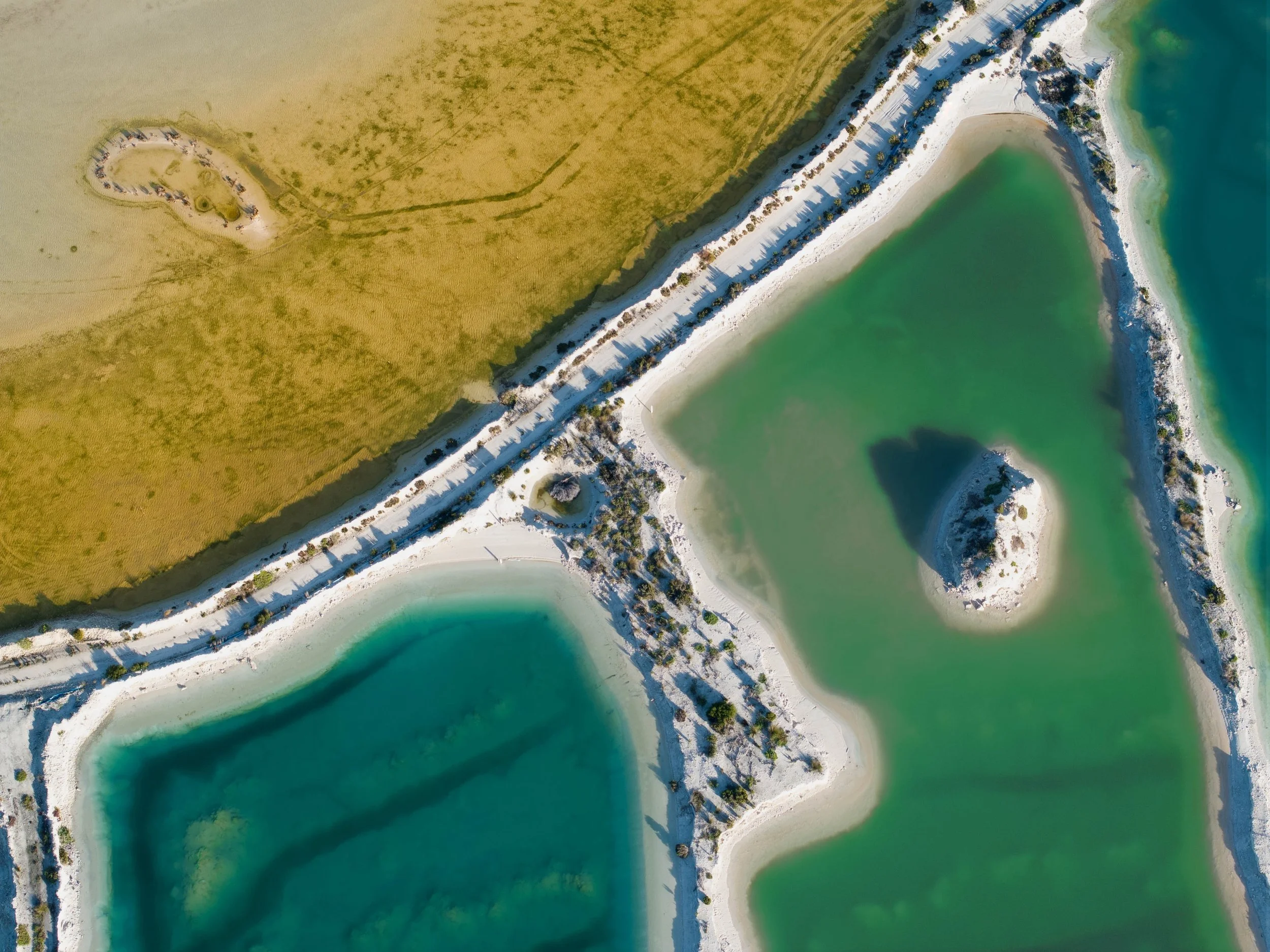The Crossing 2 — Aerial fine art photograph of white causeways crossing diagonally between turquoise and jade pools, with a small white islet casting a dramatic shadow; rich blue-green palette suited to spa, coastal, or contemporary interiors.
