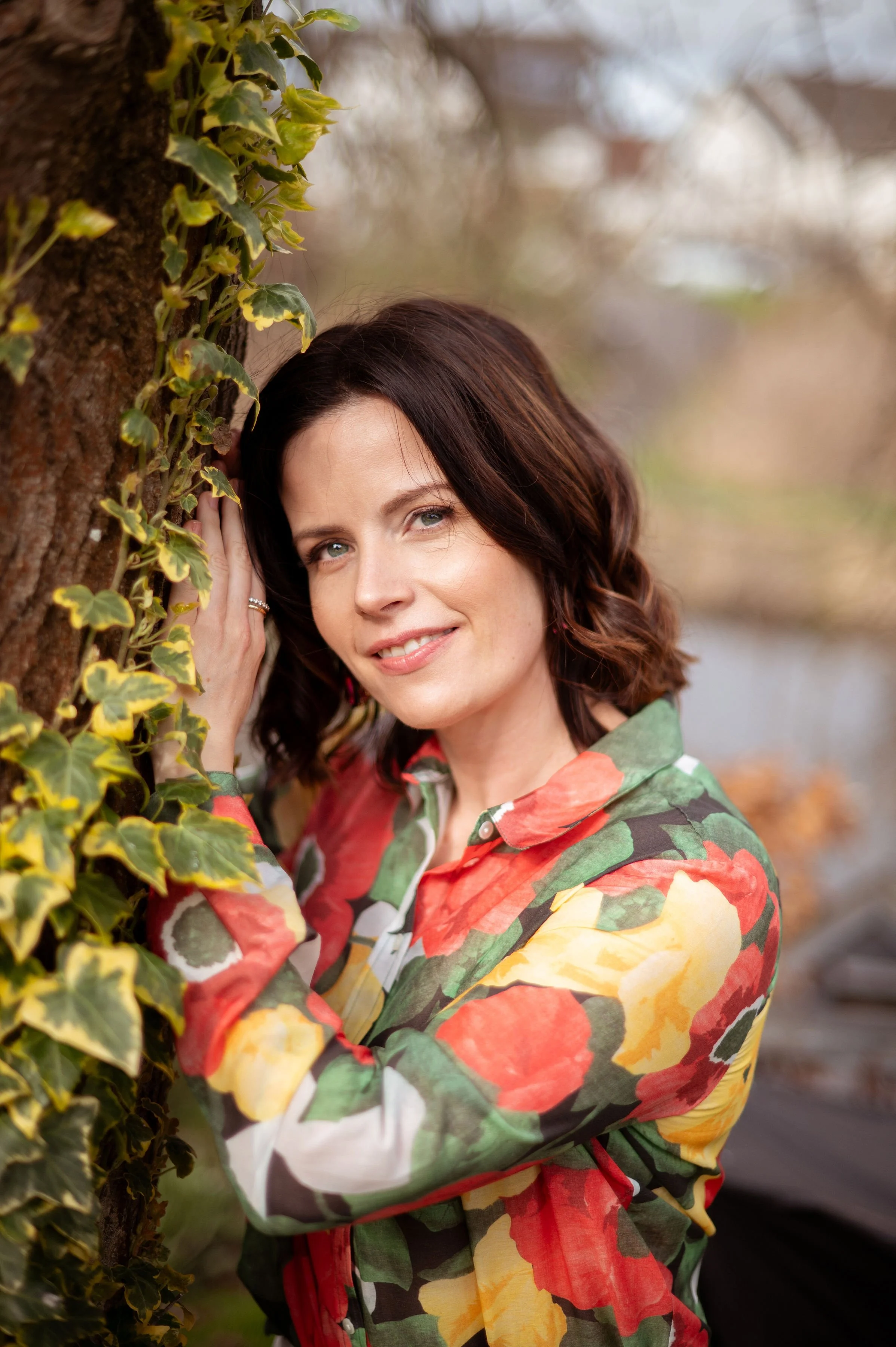 A woman with dark brown, wavy hair and green eyes leaning against a tree with ivy. She is wearing a colorful floral-patterned shirt and smiling at the camera.
