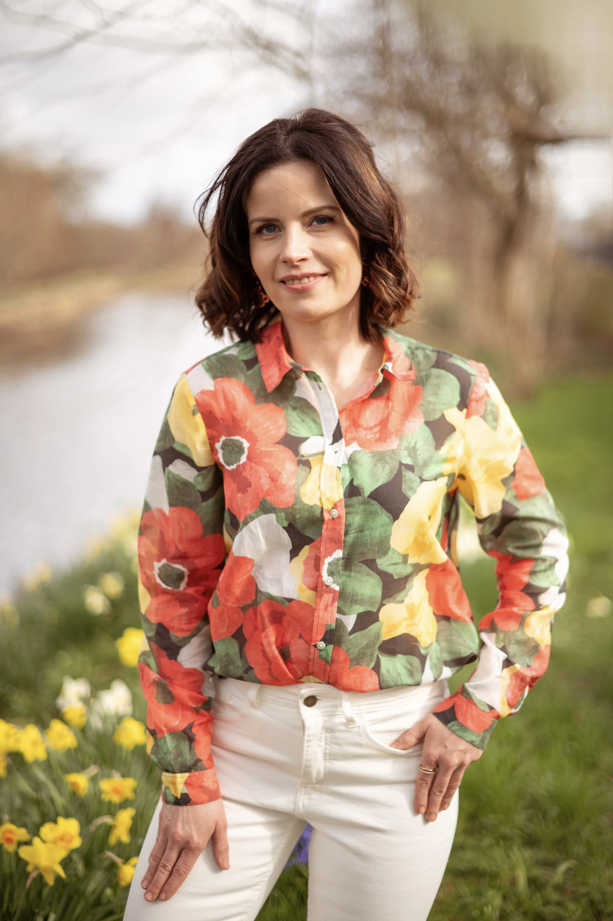 A woman with shoulder-length brown hair smiling outdoors in a spring setting with blooming daffodils and trees in the background, wearing a colorful floral shirt and white pants.