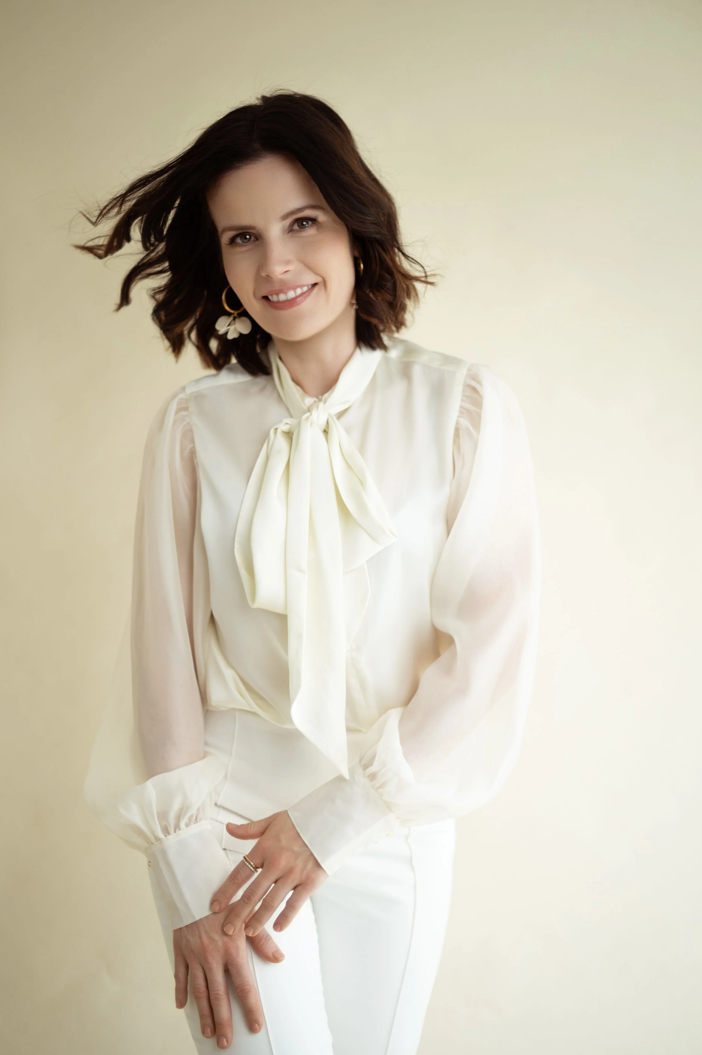 A woman with shoulder-length brown hair, wearing gold hoop earrings and floral earrings, smiling while standing against a light yellow background. She is dressed in a white blouse with a bow tie at the collar and white pants.