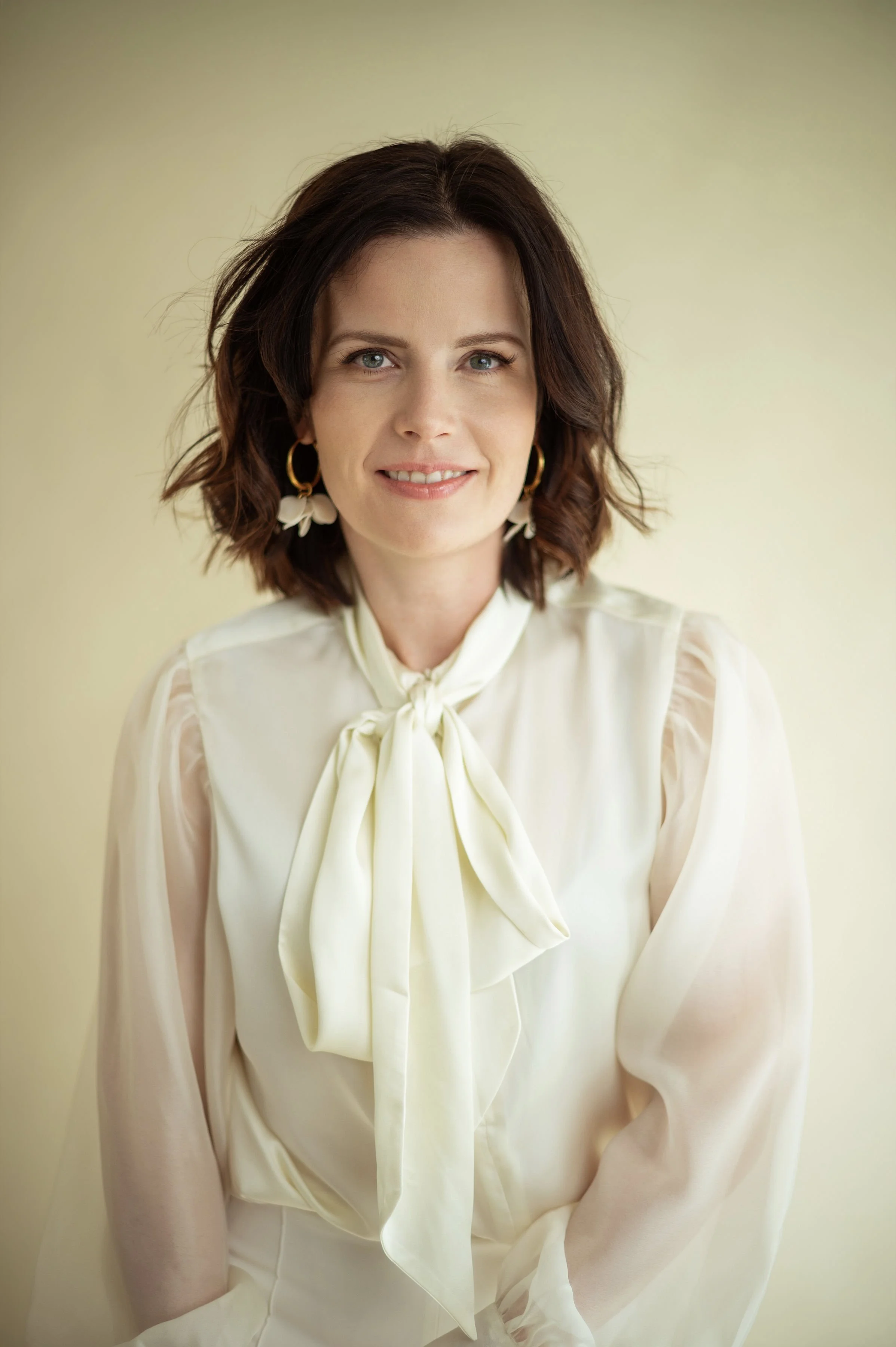 A woman with wavy brown hair, light skin, and blue eyes smiling at the camera, wearing gold hoop earrings with white flower charms, a cream-colored blouse with a tied neck bow and puffed sleeves, positioned against a plain light background.