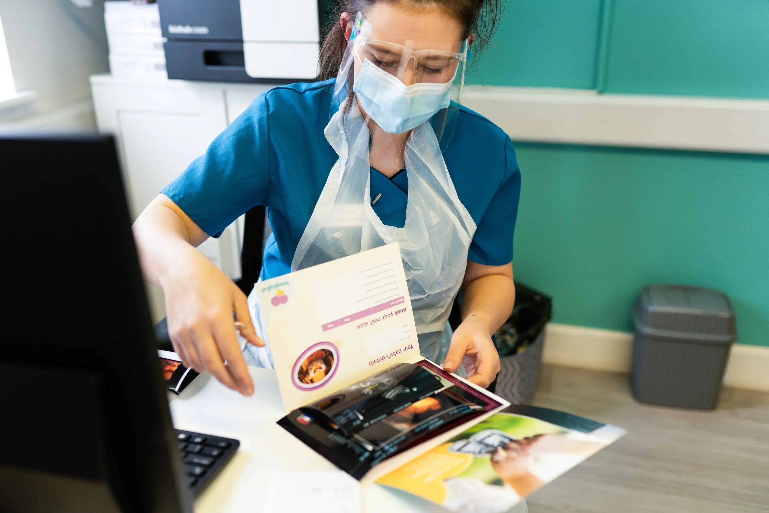 Healthcare worker in protective gear reviewing pamphlet beside computer in clinic.