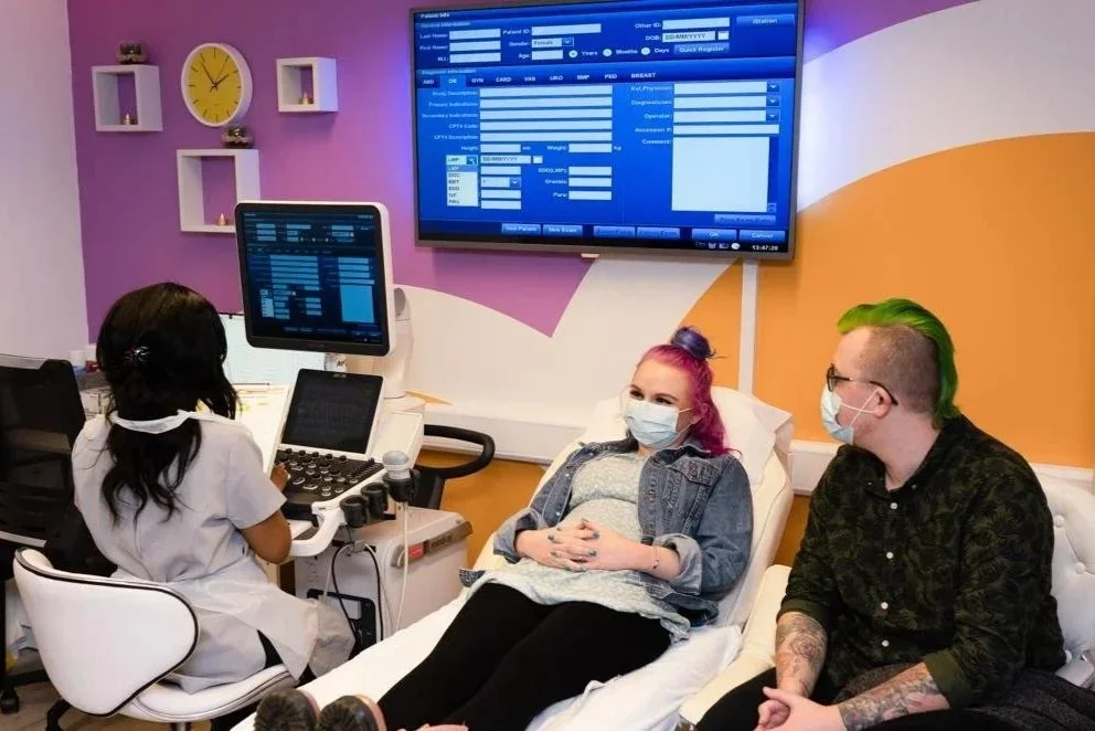 Patient lying on a bed during an ultrasound scan, with a sonographer operating the machine and a partner sitting beside her in a clinic room.